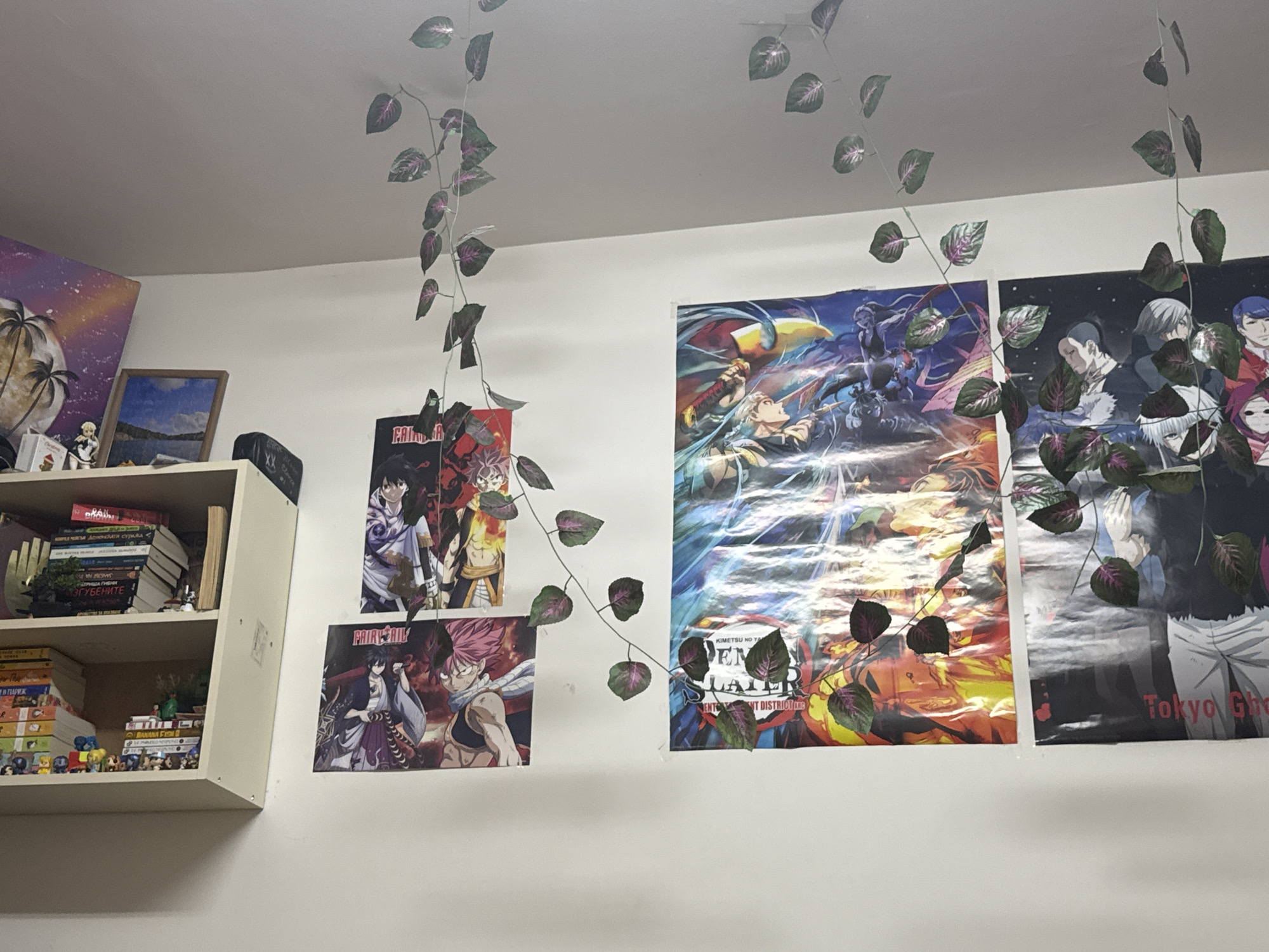 The image shows a room interior with a white wall. A white bookshelf is mounted on the left wall, filled with books, figures, and other objects. Various anime posters decorate the wall, including "Fairy Tail," "Demon Slayer," and "Tokyo Ghoul." Artificial ivy vines are hanging from the ceiling, partially obscuring the posters. The lighting appears to be from an indoor source. There is no information about the location being in Sofia, Bulgaria.