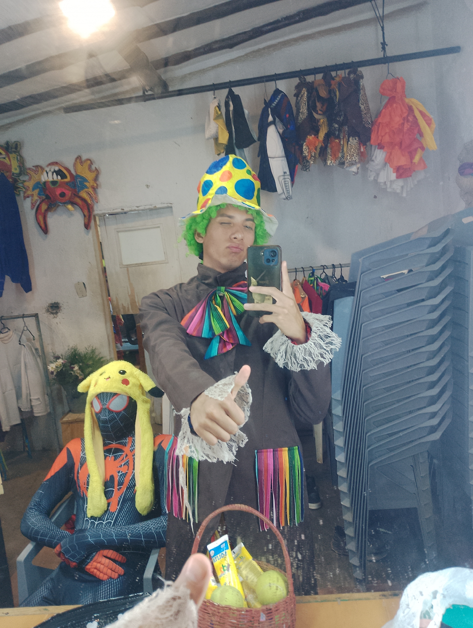 The image shows a person in a clown costume taking a selfie in what appears to be a dressing room or storage area in Porlamar, Venezuela. The individual is wearing a brown coat with a colorful ruffled collar and cuffs, a green wig, and a yellow hat with blue polka dots. The person is holding a smartphone in their left hand and making a thumbs-up gesture with their right hand. In the background, there is a Spider-Man costume with a Pikachu hat, a basket of fruit, and rows of stacked plastic chairs. Clothes and costume accessories are hanging on a rack behind the person. The room appears cluttered and somewhat disorganized. A decorative dragon head is visible on the wall.