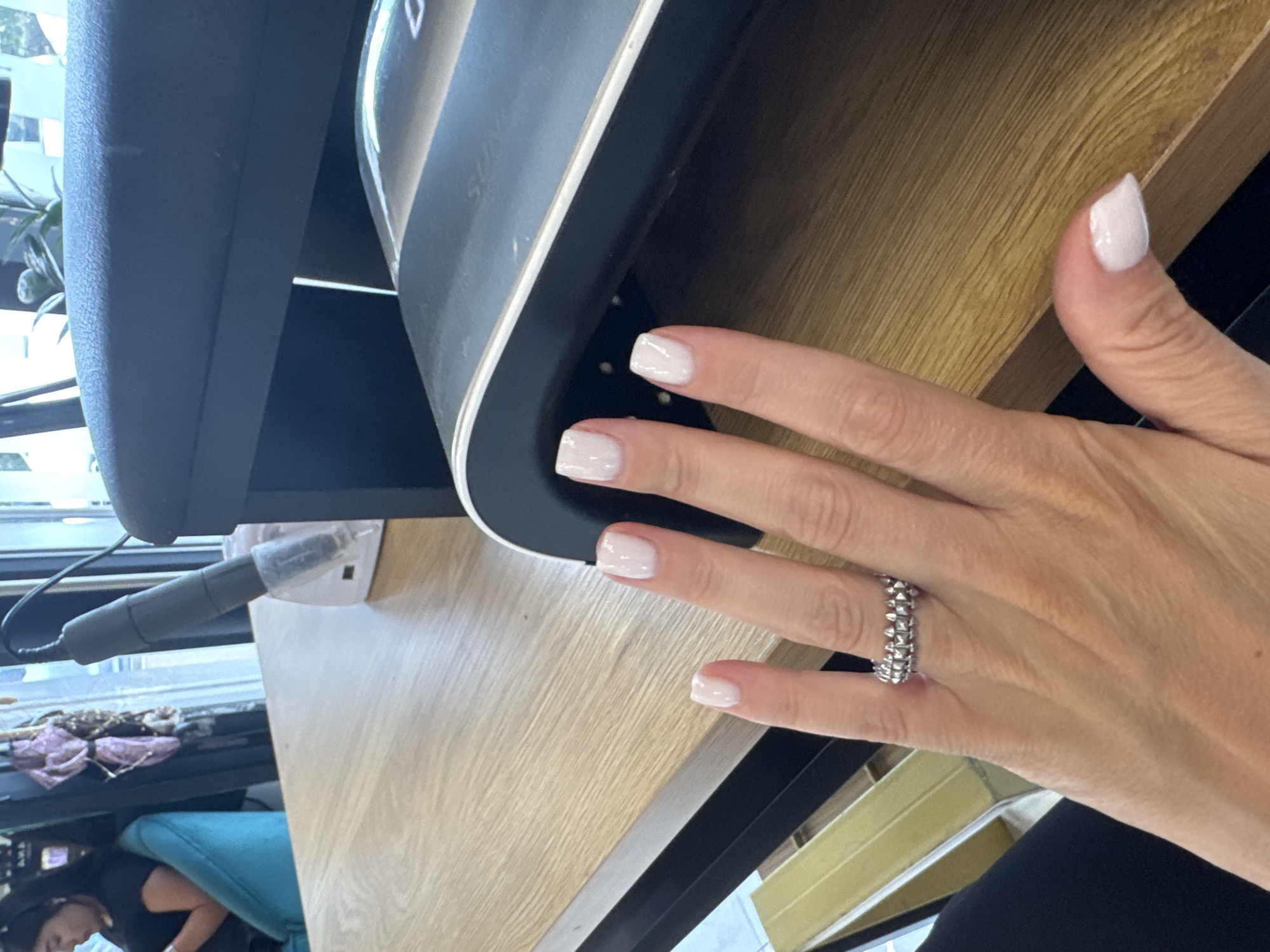 A close-up, angled shot focuses on a person's right hand with freshly manicured, white-tipped nails. The hand rests on a light-colored wooden surface, likely a desk or table. A silver, stackable ring with a textured design is worn on the ring finger. In the background, a black object, possibly a nail dryer or electronic device, is partially visible. Further in the background, a window shows a bright, daytime scene with greenery outside. The overall lighting suggests it is daytime, and the setting appears to be indoors, possibly a salon or personal workspace. There are no visible text elements in the image.