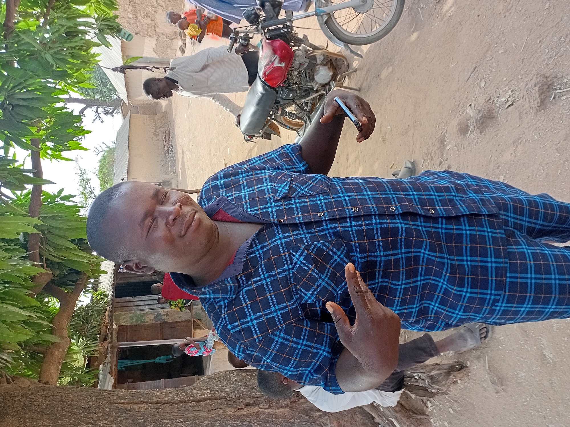 The image captures a man in Nata-Ala, Nigeria. He is wearing a blue plaid shirt and standing outside. Behind him, there's a motorcycle, another man in a white shirt, and a few buildings. A tree is on the left side of the frame. The ground appears to be dirt or sand.