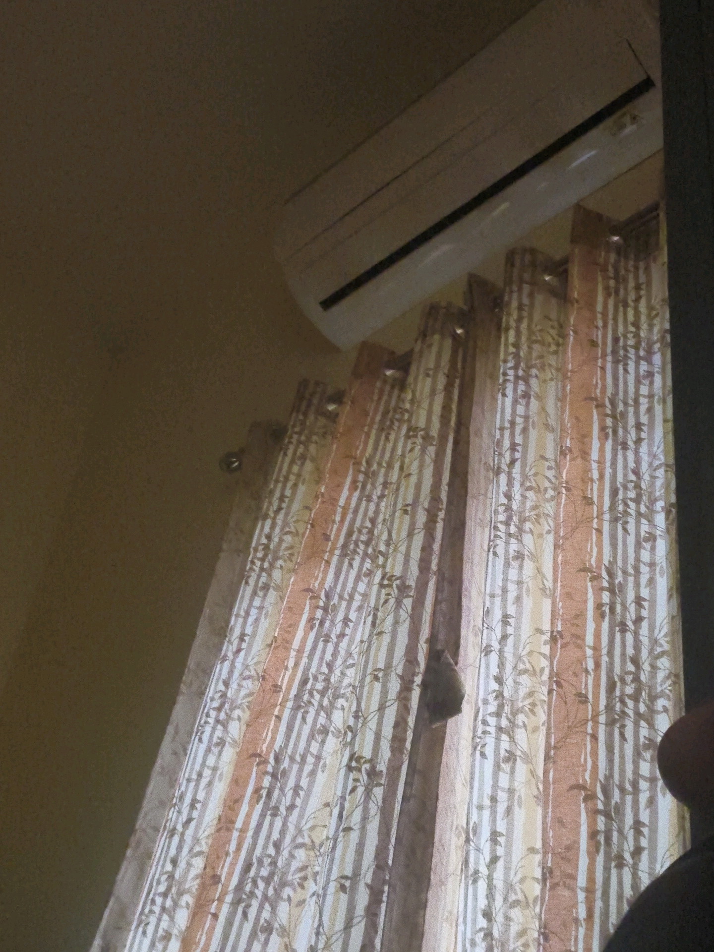 An air conditioner is mounted on a brown wall above a set of vertical striped curtains with a floral pattern. A dark, fuzzy object is hanging on the curtains to the left of the image. The scene is indoors and appears to be a room in a residential or commercial building. The image is taken from a low angle, looking upwards towards the air conditioner.