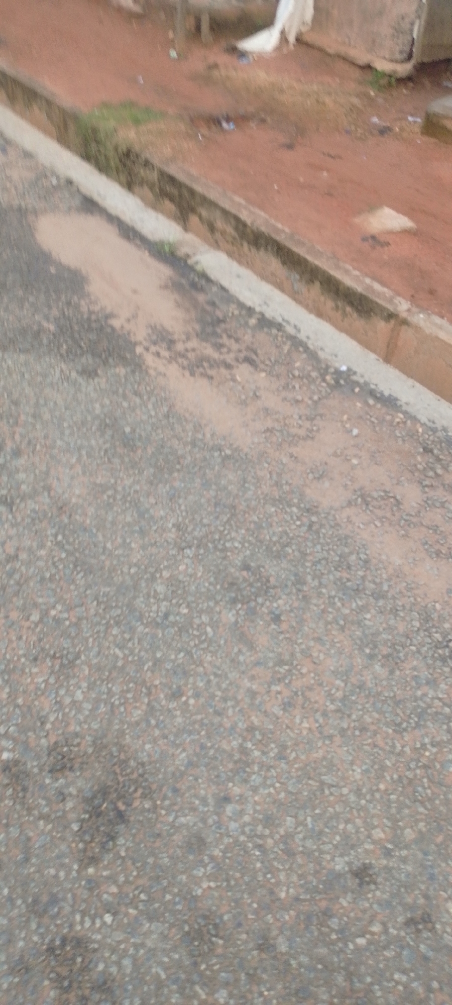 The image shows a close-up, blurry view of an asphalt road with a concrete gutter running alongside it. The road is dark gray with small stones embedded in it, and there are patches of lighter-colored dirt or sand. The gutter is lighter in color and appears to be slightly worn. Beyond the gutter, the ground is a reddish-brown color, likely dirt or clay, with some scattered debris, including what appears to be a white plastic bag. The overall scene suggests an outdoor urban or semi-urban environment, possibly a street or walkway, with the lighting indicating it could be daytime, though the blurriness makes it difficult to ascertain specific details about the surroundings or any potential human presence.