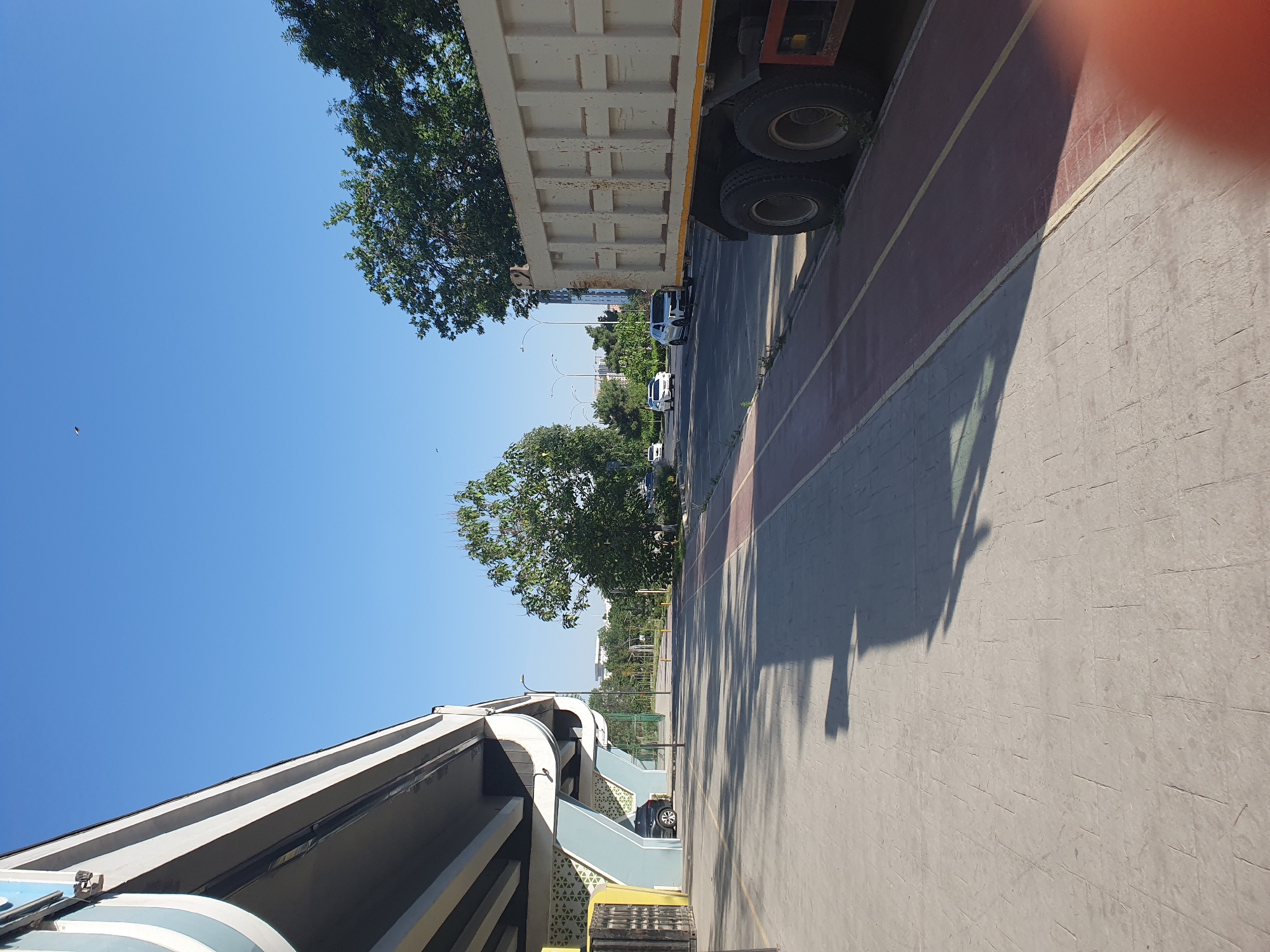 The image depicts a street scene in Tashkent, Uzbekistan, under a clear blue sky. In the foreground, to the left, is a section of an elevated highway structure with gray and white concrete supports and a curving underside. A dark-colored car is parked beneath this structure, next to a yellow gate.
To the right of the highway structure is a paved walkway with a patterned surface. Beyond this walkway is a road with a red-colored bike lane marked by yellow lines. A large, beige dump truck with dual rear wheels is parked along the edge of the bike lane, facing away from the viewer.
The road stretches into the distance, lined with trees and streetlights. Several cars, including white sedans and a silver SUV, are visible on the road. In the background, more trees and buildings can be seen, suggesting an urban environment. The overall scene is one of a sunny day in a city with infrastructure including elevated roads and designated lanes for different modes of transport.