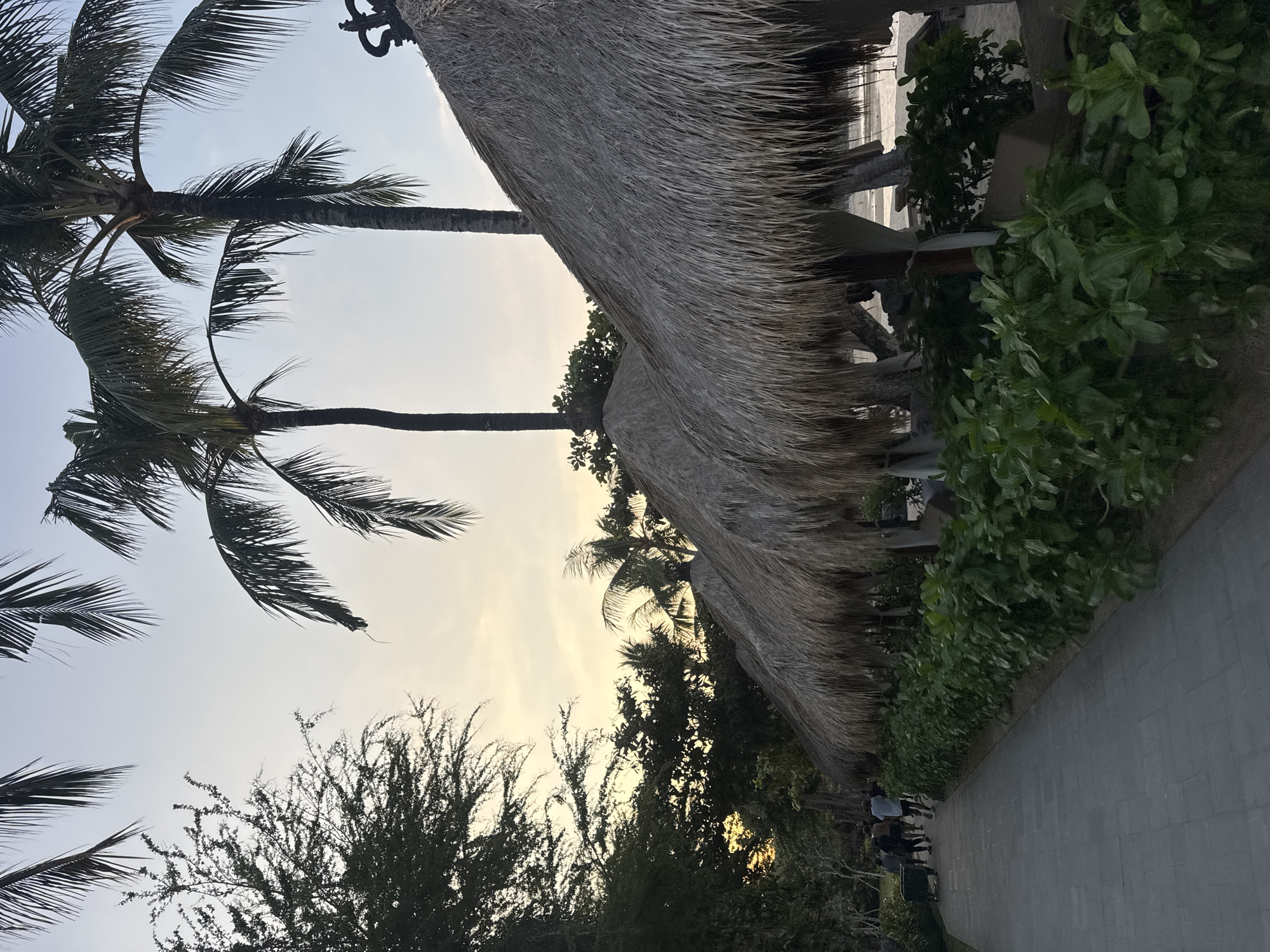 A paved pathway runs alongside a row of thatched-roof cabanas on a beach in Kuta Selatan, Indonesia. Several palm trees are visible, with their fronds silhouetted against a pale sky. A group of people are walking away from the camera down the path, some appearing to be pushing a cart. Lush green foliage lines the edge of the pathway and surrounds the cabanas. The overall scene suggests a tranquil beachside resort setting, possibly during the late afternoon or early evening.