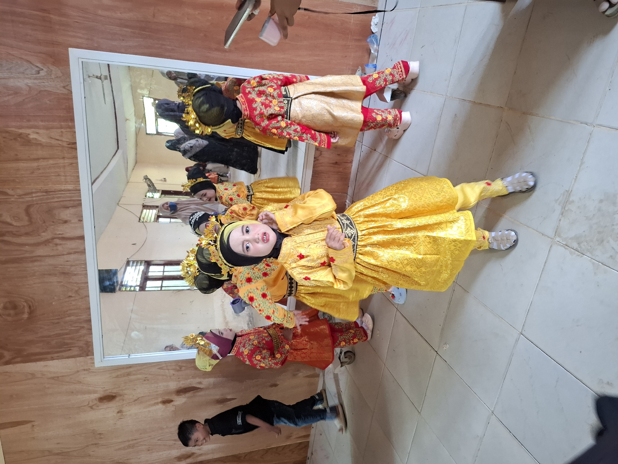 In Rhieng Blang, Indonesia, a group of young children, predominantly girls, are dressed in vibrant, traditional golden and red performance attire, complete with ornate headwear. They are positioned in a room with light-colored tiled flooring and wooden paneled walls. A large mirror dominates one wall, reflecting more children in similar costumes and several adults, some of whom appear to be supervising or assisting. One child in the foreground, wearing a bright yellow outfit, looks directly at the camera. In the background, a young boy in a black shirt and jeans stands to the left, observing the scene. The environment appears to be a preparatory space for a performance or cultural event.