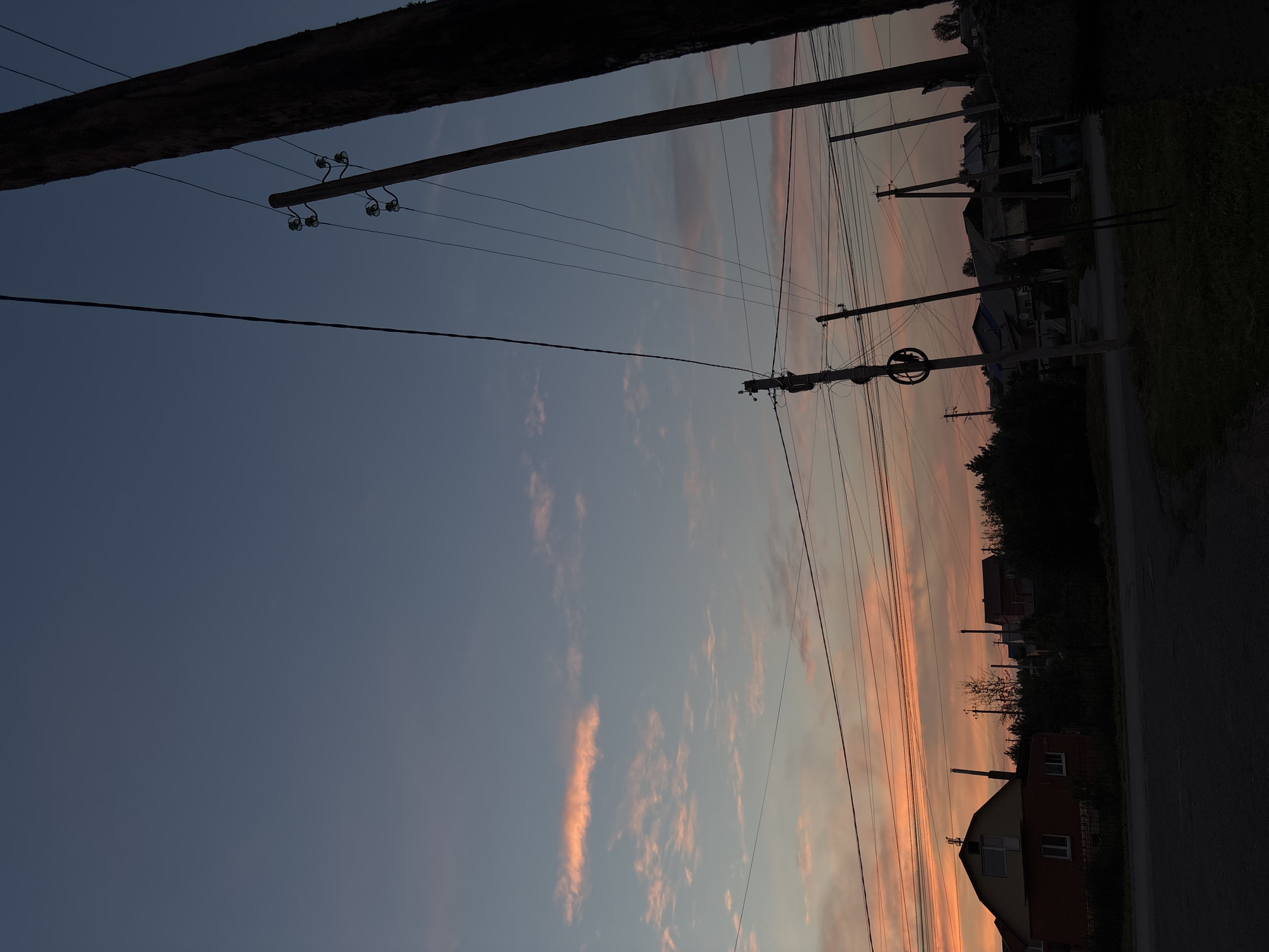 The image depicts a street scene in Vishnevka, Russia, during a sunset. The sky is a gradient of deep blue at the top, transitioning to hues of orange and pink near the horizon, with scattered clouds tinged with pink. In the foreground, a dark asphalt road curves to the left. On the right side of the road, a large, dark wooden utility pole stands prominently, angled slightly. Several other utility poles with wires are visible stretching across the frame, forming a dense network.
To the left of the road, a two-story brick house with a light-colored gable roof is visible. It has several windows and a chimney. Behind this house and stretching into the distance, a dense line of dark trees forms the backdrop. Further back and interspersed with the trees are several other residential structures, mostly silhouetted against the colorful sky. The overall atmosphere is calm and somewhat rural, characteristic of a village or small town setting at dusk. There are no people or animals visible in the image. The lighting suggests it is either sunrise or sunset, with the sun below the horizon.