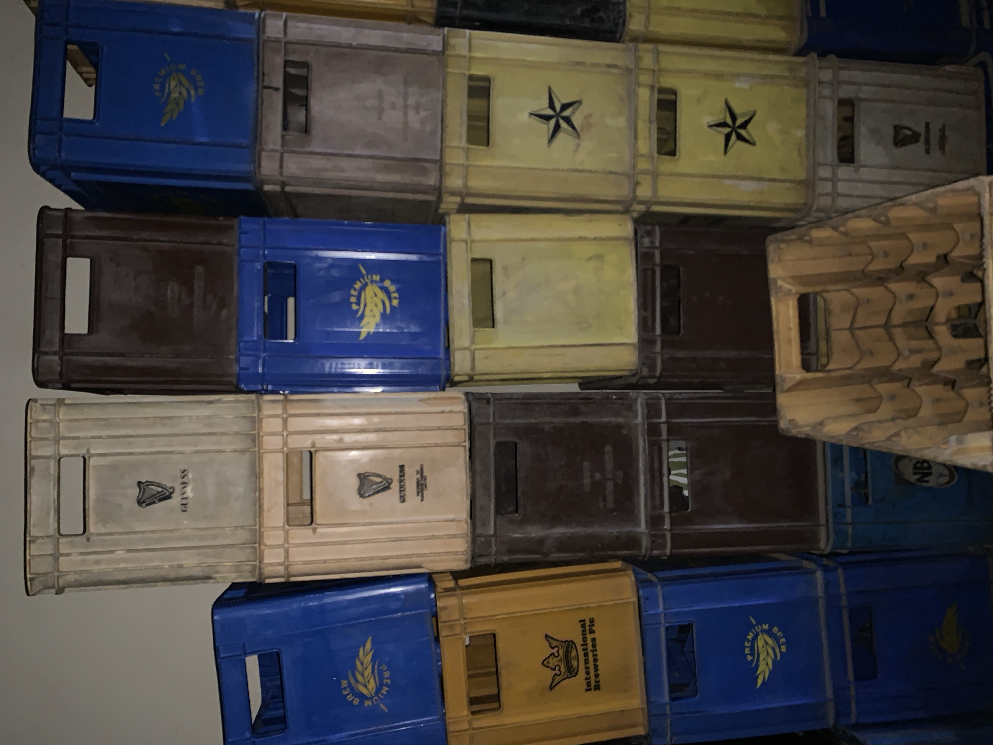 A stacked arrangement of plastic beer crates dominates the scene. The crates are a mix of colors including blue, brown, yellow, and off-white. Various brewery logos and emblems adorn the sides of the crates, including "Premium Brew," a harp-like symbol labeled "Guinness," a five-pointed star, and "International Breweries Plc." One crate is oriented at an angle. The location in the city of New Stadium, Nigeria is not discernible from the image.