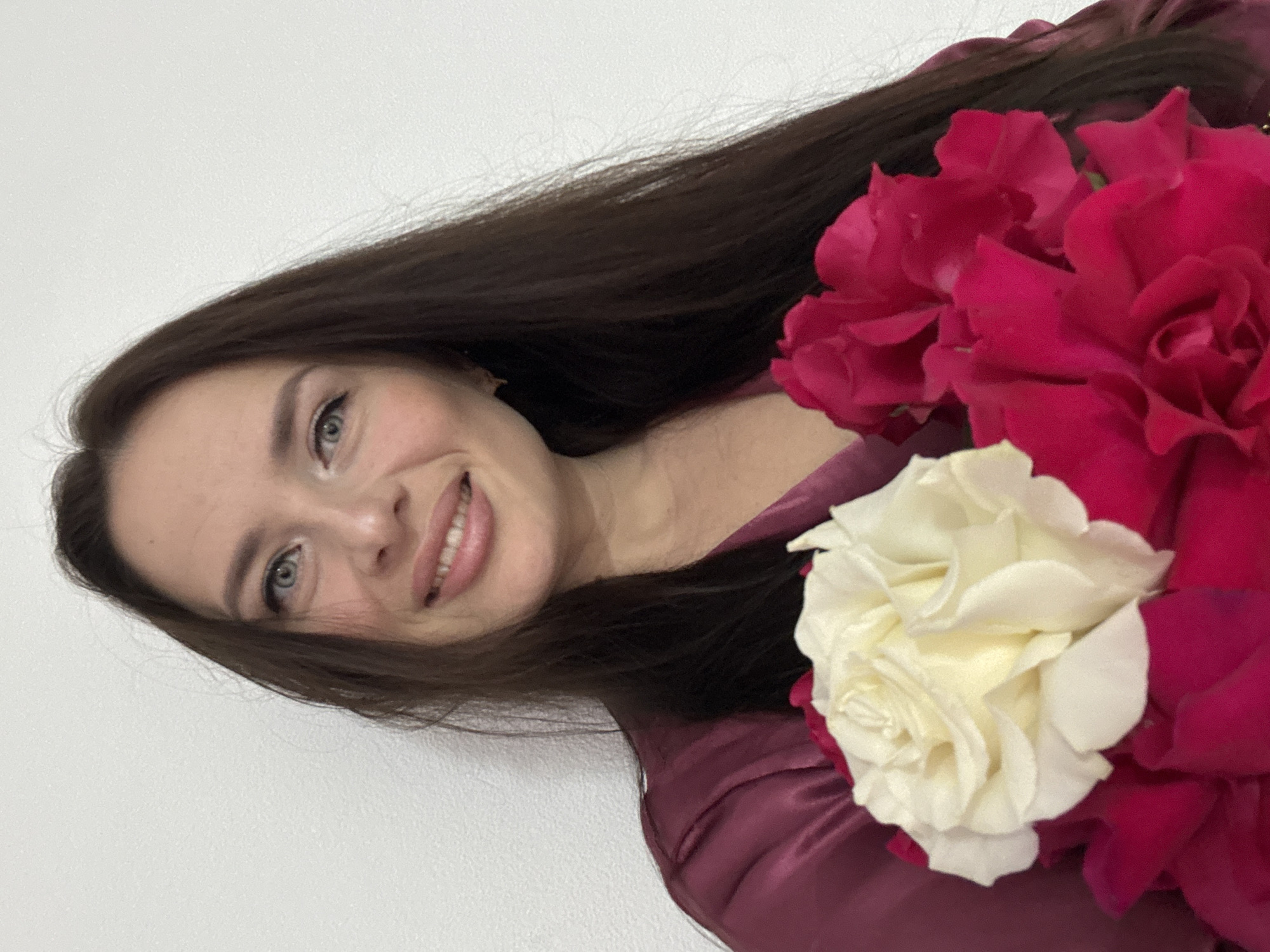 A medium shot shows a woman with long, dark brown hair and blue eyes against a plain white wall. She is smiling and looking at the camera. She is wearing a dark pink satin robe. She is holding a bouquet of roses, with white and dark pink blooms visible. The setting is not specified to be in Rusalevka, Russia.