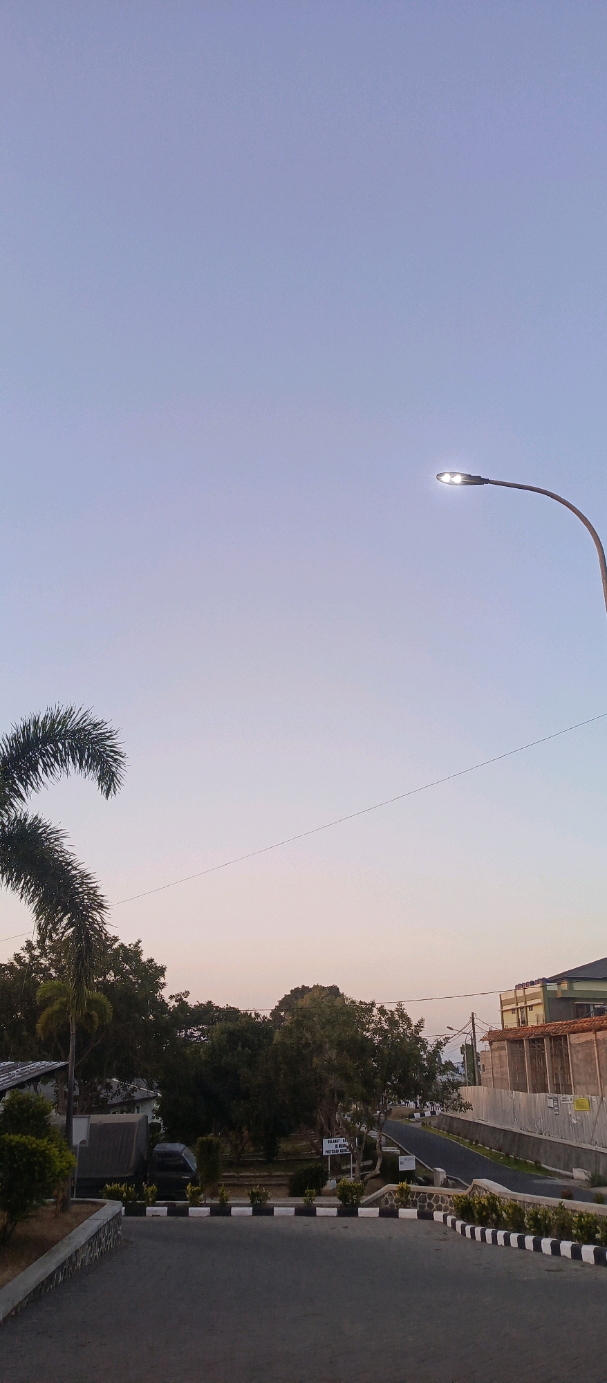 The image depicts a serene outdoor scene at dusk, possibly in Batu, Indonesia. A wide asphalt road with black and white striped curbing curves through the foreground. On the left, a palm tree's fronds extend into the upper left corner. Beyond the road, a variety of trees form a dense line, interspersed with a few buildings. One building, partially visible on the far left, appears to be a single-story structure with a gray roof. Further to the right, a multi-story building with a tiled roof and light-colored walls is visible, bordered by a construction fence. A single street light with two illuminated bulbs stands tall on a curved pole in the upper right quadrant of the sky, casting a warm glow. The sky transitions from a pale blue at the top to a soft, warm orange and pink hue near the horizon, indicating the approaching night. The overall atmosphere is tranquil and peaceful, with no people or animals visible in the frame.