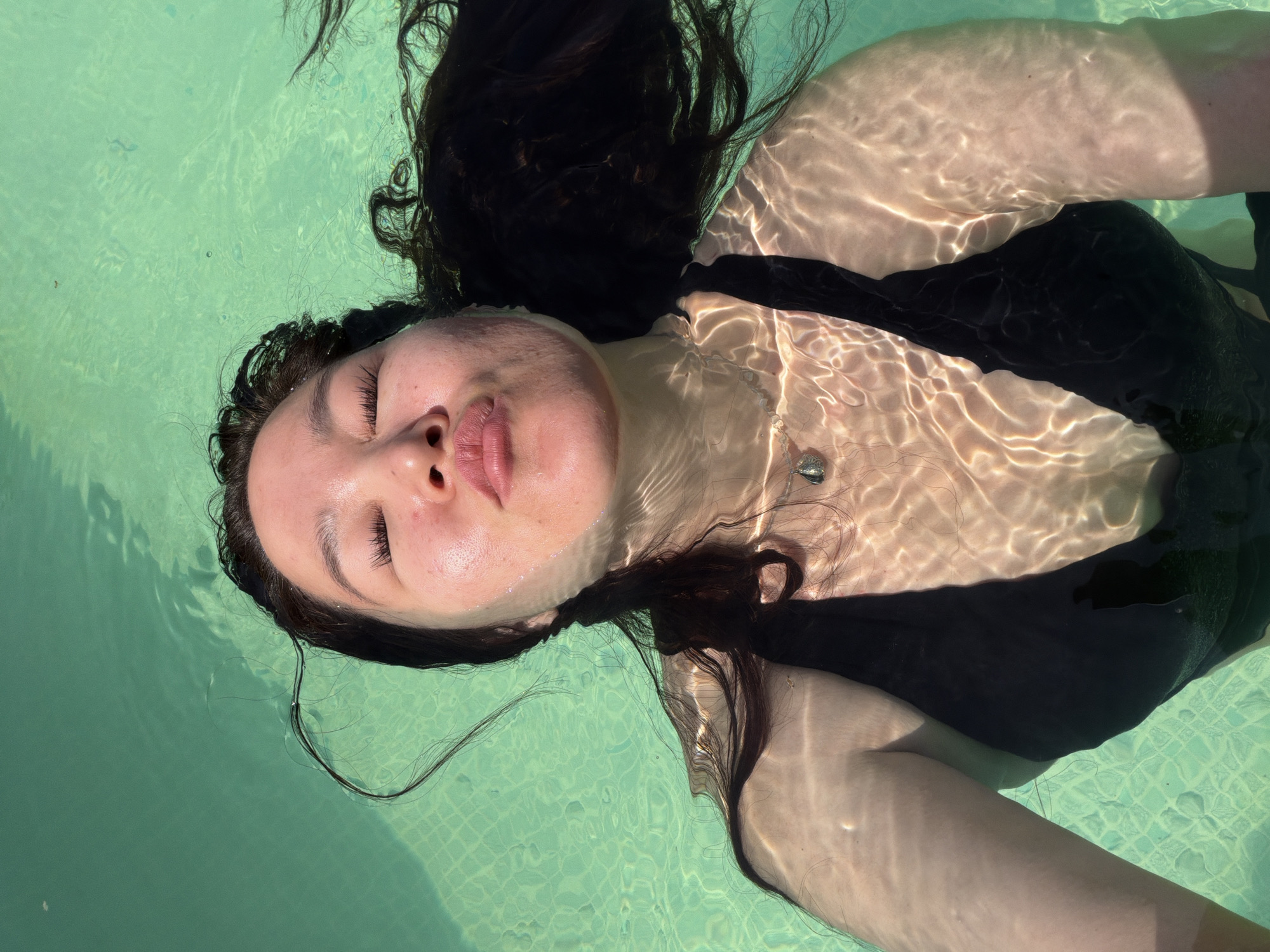 A woman with closed eyes floats on her back in a pool. The pool's water is light green, and sunlight casts rippling patterns on her skin and the pool's surface. The woman has dark, wet hair spread around her head in the water. She is wearing a black swimsuit. The location appears to be in Unknown, Unknown.