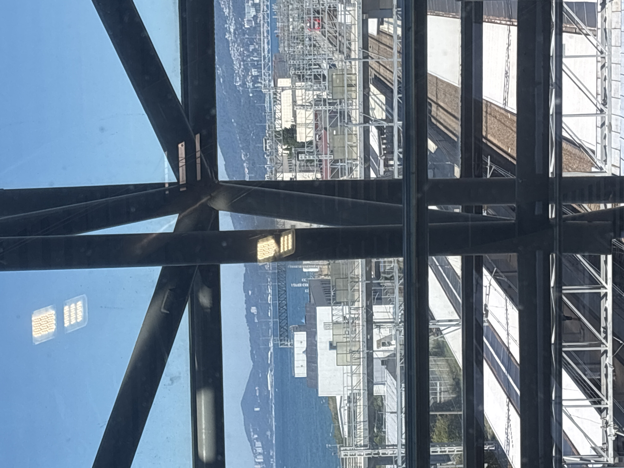 The image provides a view through a window with structural supports, looking out over a city with a body of water and mountains in the distance. In the foreground, vertical and diagonal dark metal beams dissect the view. Through the beams, we can see a bright blue sky.
The middle ground of the image reveals a cityscape with a body of water, likely the Black Sea, bordering a coastal area. Buildings of various sizes and architectural styles are visible, including white structures that appear to be industrial or commercial. Train tracks and platforms are also prominent, with several covered platforms suggesting a train station or depot. One red train car is visible on the tracks.
The background features rolling hills or mountains covered in vegetation and dotted with buildings, indicating the city extends inland. The overall impression is a view of a coastal city with significant infrastructure. The specific location is the Sochi train station in Sochi, Russia, which is known for its coastal setting and proximity to mountains. The reflection on the glass shows what appear to be illuminated numbers on a rectangular surface, possibly related to the building's interior lighting or signage.