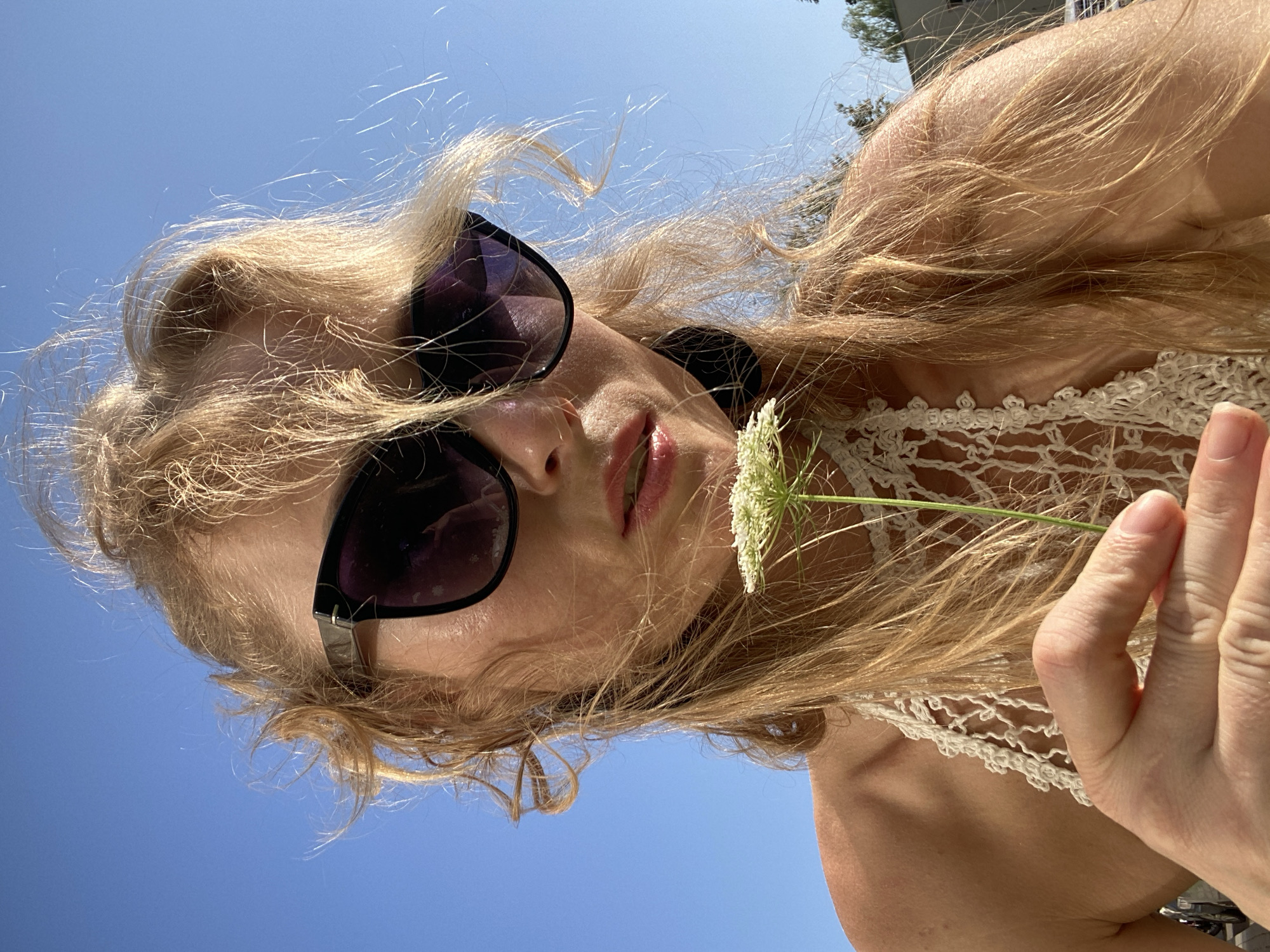 A woman with blonde hair and sunglasses is holding a white flower in front of her face, looking at the camera. She is wearing a white crocheted top. The background is a clear blue sky with some trees visible in the distance. The image appears to be taken outdoors on a sunny day. There is no visible indication of the location being Lida, Belarus.