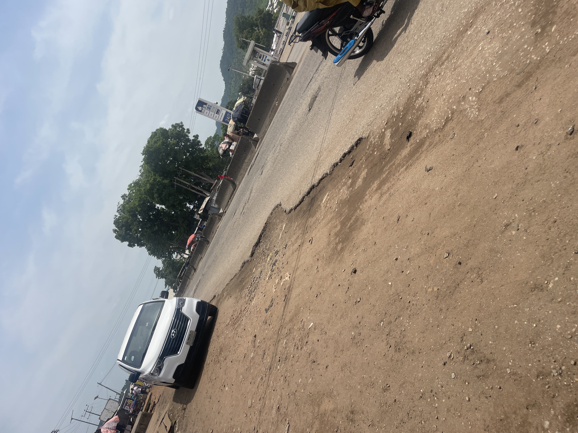 The image depicts a street scene in Lokoja, Nigeria. In the foreground, a white Hyundai Staria minivan is parked on a dirt shoulder next to a damaged asphalt road. The road exhibits significant cracks and potholes, indicating poor maintenance. Further down the road, a motorcycle with a rider is visible, heading away from the camera. Several makeshift stalls with vendors and goods are set up along the roadside, suggesting a busy commercial area. A tall sign with the "NNPC" logo, indicating a Nigerian National Petroleum Corporation station, is prominent in the background, along with a gas station. Trees and hills form the backdrop under a partly cloudy sky. The overall impression is of a bustling, potentially underserved urban environment with active commerce and transportation.