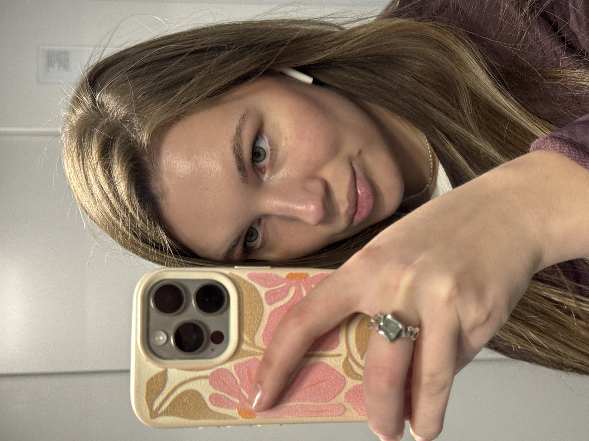A young woman is captured in a close-up, angled selfie, taken indoors. Her long, light brown hair is styled to one side, and she wears a dark purple top. She looks directly at the camera with a gentle smile, her light green eyes and softly pink lips prominent. An AirPods earbud is visible in her left ear.
Her right hand holds a smartphone, partially obscuring her face, with the back of the phone and its triple camera lenses clearly visible. The phone is encased in a cream-colored cover adorned with a distinctive pattern of large pink and tan flowers. On her ring finger, she wears a silver-toned ring featuring a rhomboid-shaped, greenish stone. A thin silver necklace is also visible.
The background consists of a plain, light-colored wall, indicating an indoor setting with bright, even lighting, suggesting daytime. A blurry, illegible white label with black text is faintly visible on the wall in the upper left. This scene is set within Vozdovac Urban Municipality, Serbia.