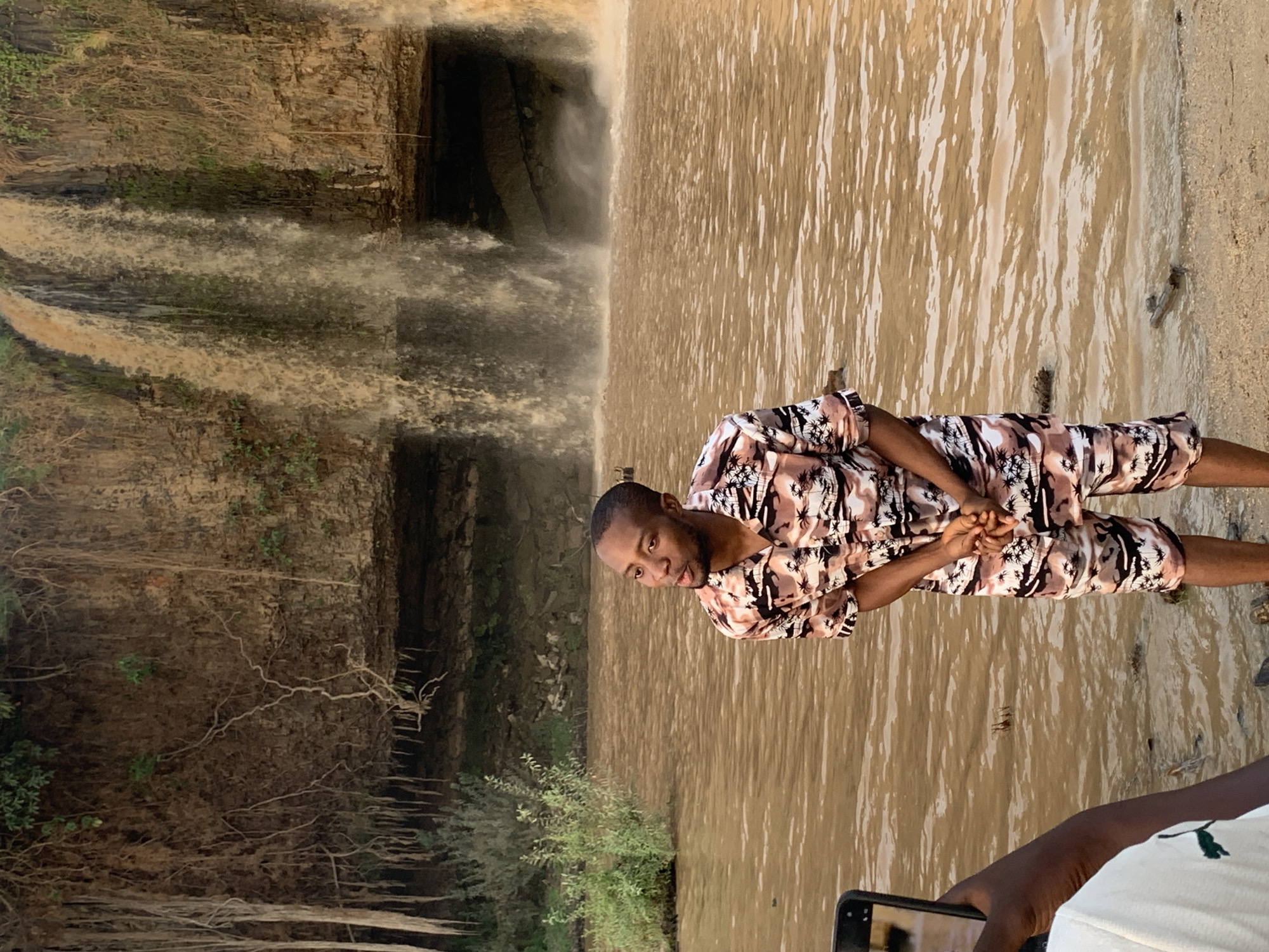 A medium-shot of a man in a brown, white, and black patterned short-sleeved shirt and shorts standing at the base of a waterfall. He is slightly turned to the left. His hands are folded in front of him. Behind him is a waterfall, and he is standing in water. The scene is in Kukum, Nigeria. A person holding a phone is visible in the lower right of the frame.