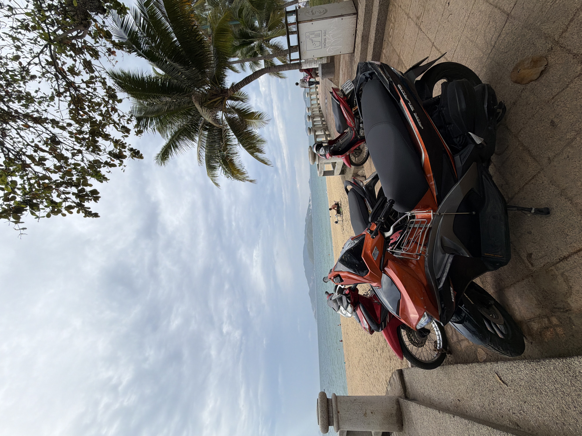 In Nha Trang, Vietnam, a scenic beachfront promenade is depicted under a cloudy sky. The foreground is dominated by parked motorcycles. In the center, an orange and black scooter, labeled "AIR BLADE", is prominently featured. To its left, a red scooter is visible, partially obscured. Further back, another red scooter is parked near a stone railing. Beyond the promenade, a sandy beach stretches towards the turquoise ocean, with a distant hilly coastline. A few individuals are scattered across the beach and the promenade, one person walking near a trash can labeled "THÙNG RÁC". Palm trees and foliage frame the upper right portion of the image, adding to the tropical atmosphere. The overall scene suggests a peaceful, overcast day at the beach.