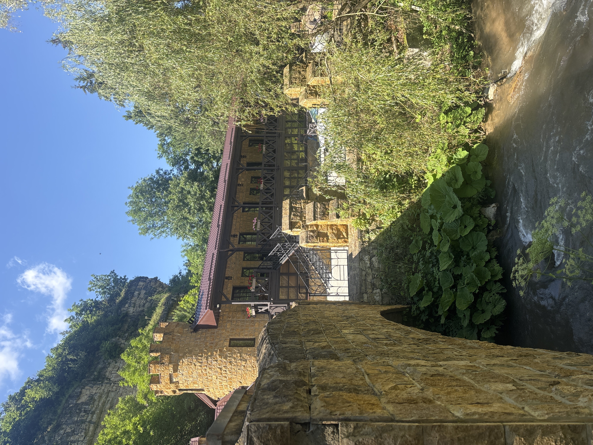 The image depicts a scenic view of a stone bridge, a building resembling a castle tower, and a multi-story structure with a balcony, all situated near a flowing river and surrounded by lush greenery and a rocky cliff face. The architecture suggests a historical or themed establishment. A small waterfall or rapids are visible in the river. The sky is clear and blue. The setting appears to be a park or resort area. This location is identified as the "Dacha of the Narzan Gallery" or "Dacha of the Mayor" in Kislovodsk, Russia, a resort city known for its mineral springs. One person is visible on the lower balcony of the multi-story building.