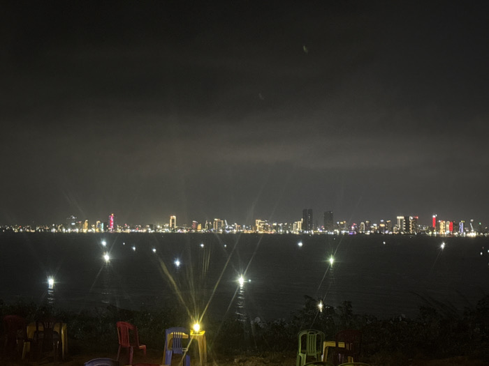 The scene is a nighttime panorama of the Da Nang city skyline viewed from Sơn Trà Ward, Vietnam. The foreground features dark ground cover and several plastic chairs of red, blue, and green coloration. Two yellow-glowing lanterns are visible on the ground, producing prominent starburst lens flares. The midground consists of a dark body of water, likely a bay or river, reflecting numerous bright, starburst-shaped lights. The background displays a densely illuminated city skyline. High-rise buildings with varying light patterns are visible, including a central skyscraper emitting a distinct red illumination and others featuring white, yellow, and blue lights. The sky is dark and overcast, showing no celestial bodies. Faint, elongated light streaks are present in the upper-center sky. No individuals are visible.