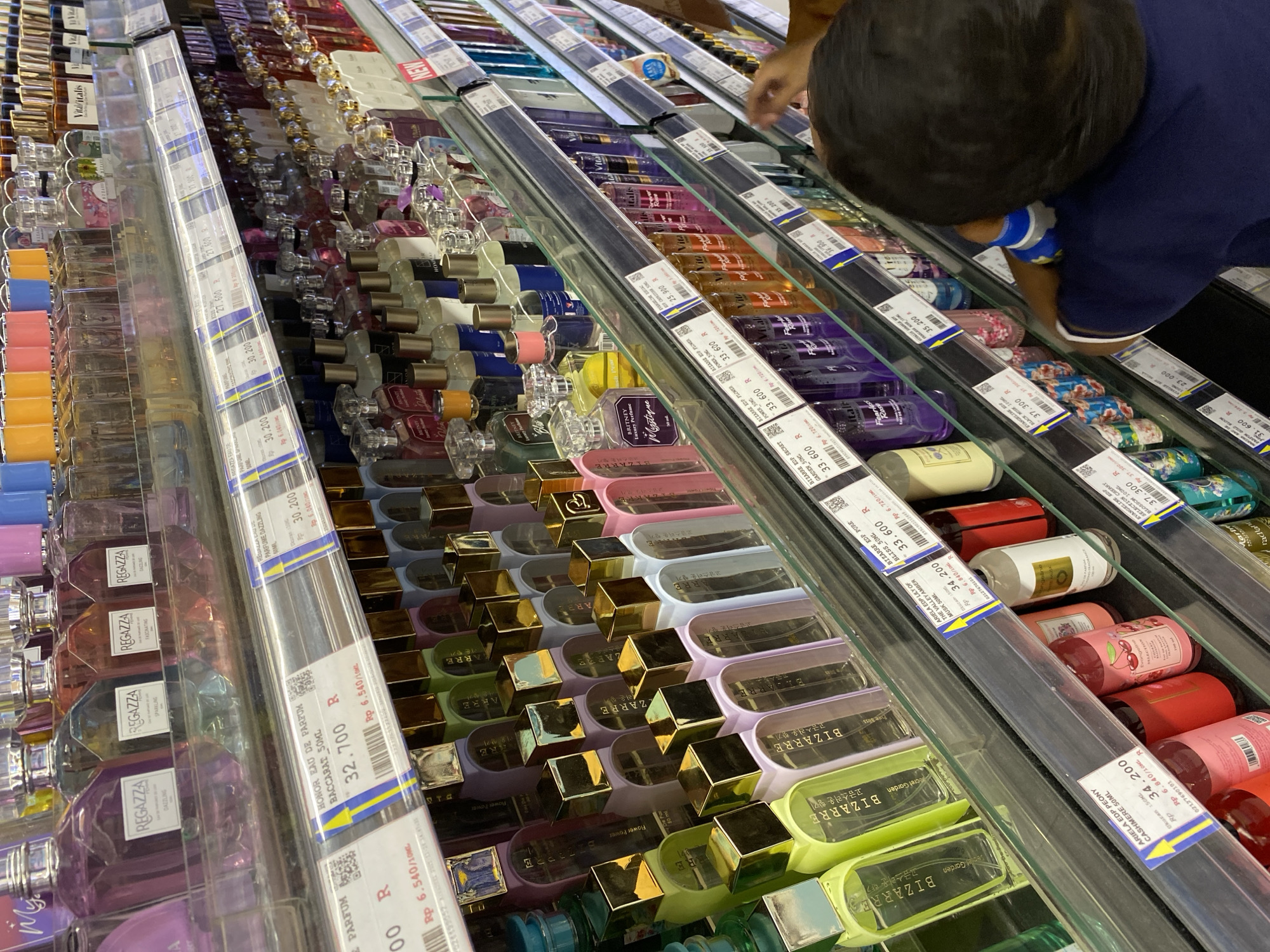 This image shows a well-lit retail store, likely a department store or supermarket, displaying a wide variety of perfumes on multiple shelves. The products are primarily bottled perfumes of various shapes, sizes, and colors. Many bottles have gold-colored caps or accents. Product labels with prices are visible on some shelves, indicating a commercial setting. Several price tags show prices in Indonesian Rupiah (Rp). The overall arrangement suggests a perfume or cosmetics section of a store. There is a person, a young child with dark hair, in the foreground, facing away from the camera and looking at the merchandise. The environment appears clean and organized. The image was taken in Banda Aceh, Indonesia.
