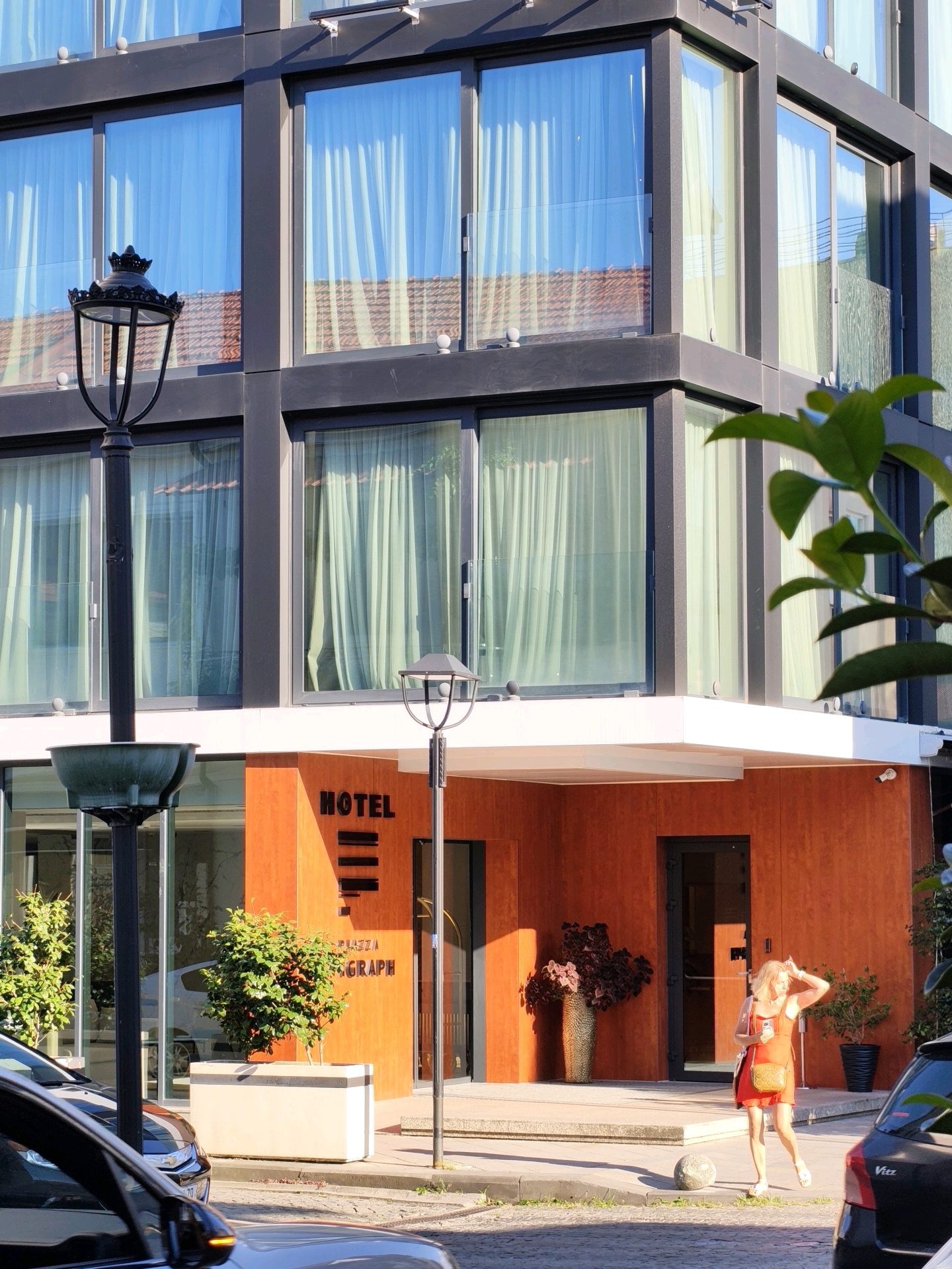 The image captures a sunny afternoon scene outside a modern hotel in Batumi, Georgia. The hotel features a sleek facade with large, reflective windows and an orange-colored lower level. A woman in a red dress is standing on the sidewalk, looking at her phone, and appears to be adjusting her hair. A black car is parked in the foreground. In the background, a planter with a lush green bush sits outside the hotel entrance, and decorative vases are visible. The architecture suggests a contemporary design with clean lines and a mix of materials. The lighting indicates a bright day, with shadows cast on the ground.