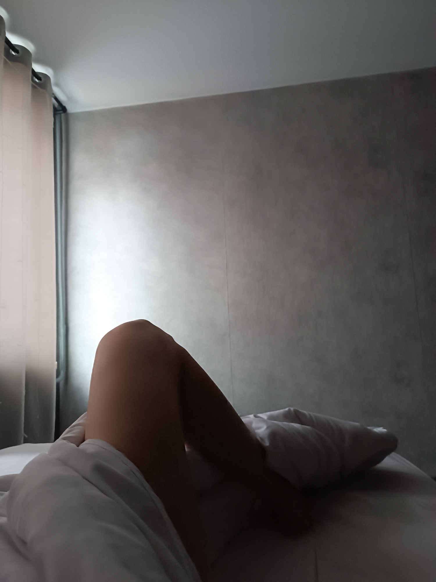 A low-angle, medium shot shows a partially covered person in bed in a room with a gray wall. A white sheet covers the person's lower body, and one leg, bent at the knee, is visible. Light comes from a window covered by tan curtains. The location is unknown.