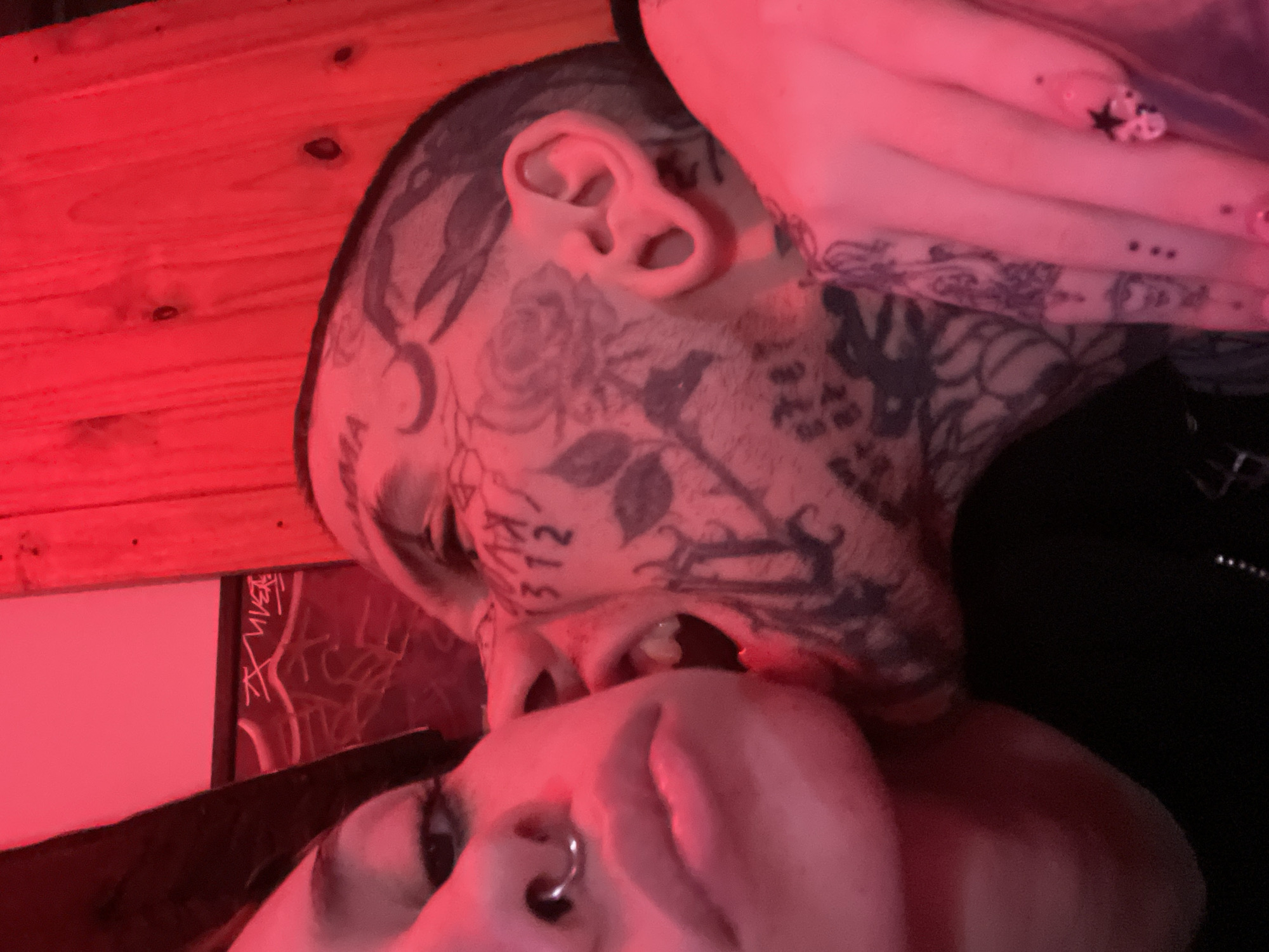 The image portrays an intimate, close-up scene featuring two individuals, heavily illuminated by a striking red light. The setting appears to be indoors, dimly lit, likely at night.
In the foreground, a person with fair skin is partially visible, featuring a nose ring. Their head is angled, showing an eye and part of their face, directed towards the other individual.
The central subject, positioned above the first, is extensively tattooed across their head, face, and neck. Notable tattoo designs include a crescent moon, roses, other floral and foliage patterns, and the distinct numbers "372" along with a partial "M" on their cheek. This person's ear shows visible modifications. Their mouth is open, revealing teeth, suggesting an intense interaction such as kissing, a playful bite, or very close communication with the person below. A hand, likely belonging to this tattooed individual, is visible in the upper right, adorned with painted nails and a star-shaped embellishment.
The close proximity and expressions of the subjects convey a sense of intimacy or strong emotional connection. The artificial red light casts a warm, dramatic, and somewhat mysterious ambiance over the entire scene. In the background, wooden planks, possibly a wall or furniture, are bathed in the same red glow. To the left, a framed item, potentially a piece of art or paper, displays faint, stylized white markings resembling graffiti, with some illegible text. No specific visual cues within the image confirm the location as Buenos Aires, Argentina, though this context is provided.