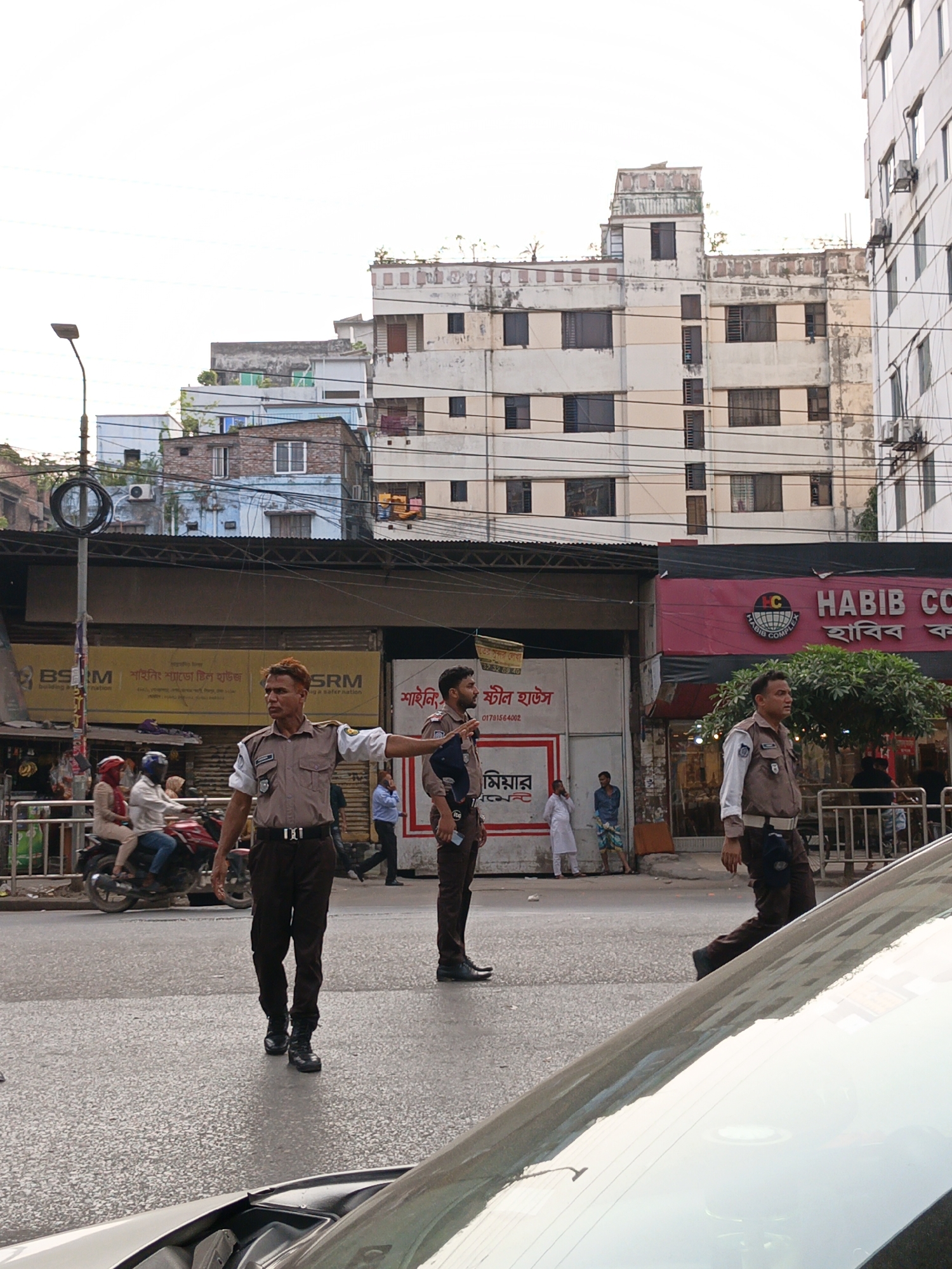 The image captures a street scene in Dhaka, Bangladesh, featuring three uniformed traffic police officers. One officer is directing traffic with an outstretched arm, while another stands nearby. The third officer walks in the opposite direction. A motorcycle with two passengers is seen to the left of the officer directing traffic. Behind the officers are various buildings, including one with the sign "HABIB COMPLEX". A sign for "SRM" is also visible. The foreground includes the hood of a car. The background shows a densely built environment with multiple buildings stacked behind each other. The time of day appears to be daylight.