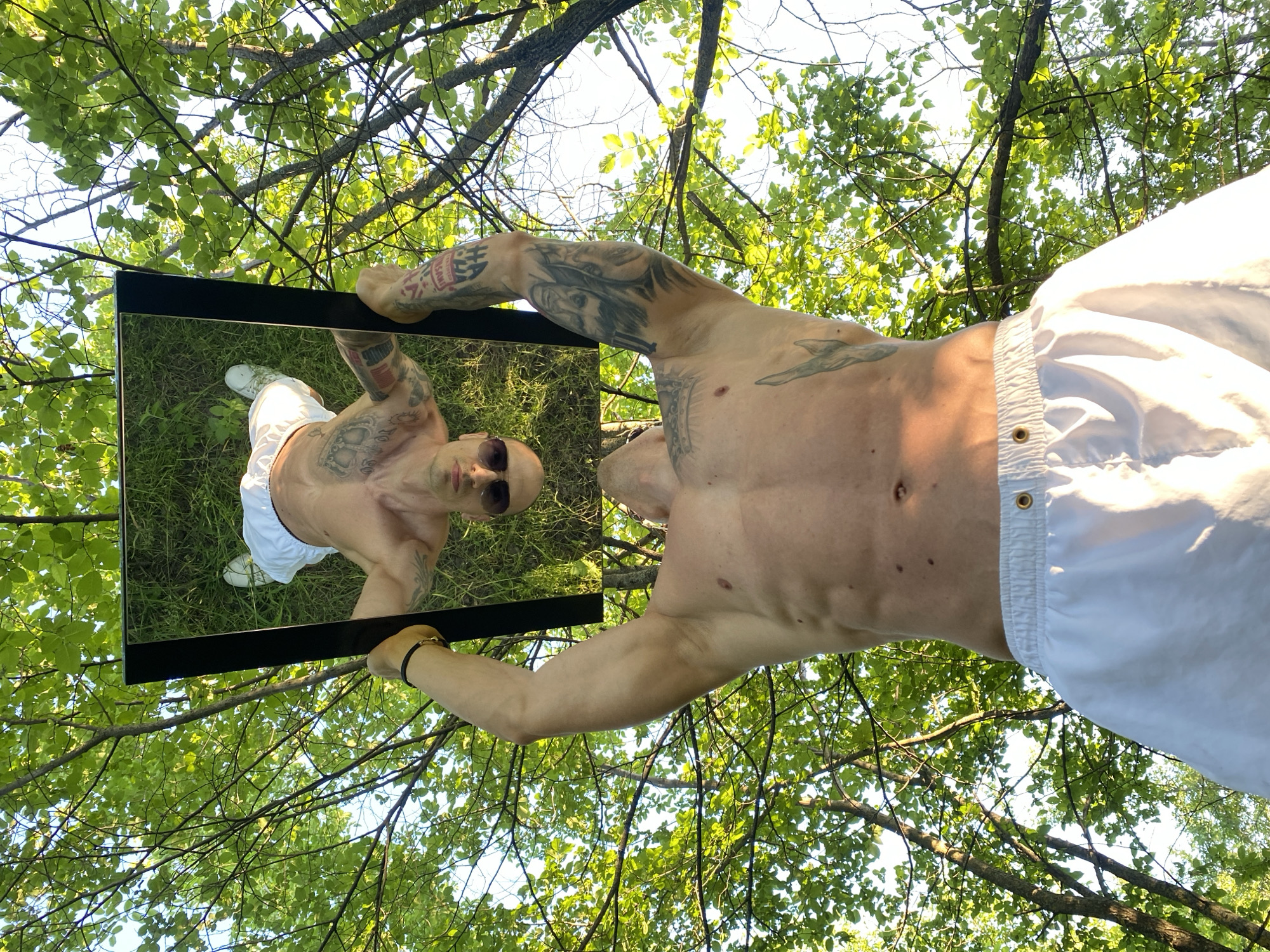 A shirtless man with tattoos is holding a rectangular mirror outdoors, reflecting his image against a backdrop of tree branches and leaves in bright sunlight. He is wearing white shorts and sunglasses, and the mirror shows him standing on grassy ground with white sneakers. The location is not discernible to be in Pleven, Bulgaria from the image.