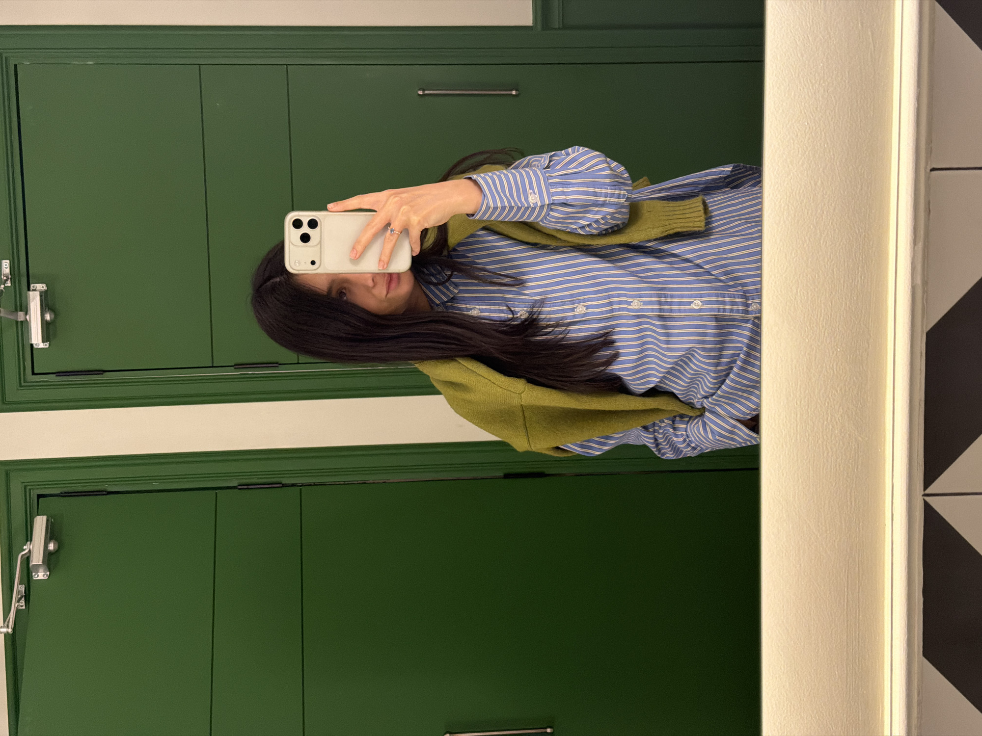 A mirror selfie depicts a female in a public restroom setting, identifiable as The Alexander, a Luxury Collection Hotel, Yerevan, Armenia, specifically within its public restroom facilities at Abovyan St 2/1. The subject, a woman with long dark hair, is visible from the upper torso, wearing a blue and white vertical striped button-down shirt with a lime green knitted sweater draped over her shoulders. She holds a light-colored smartphone with a triple-lens camera in her right hand, angled to capture her reflection. A ring is present on her left ring finger. Her left hand is partially visible, appearing to be in a pocket. The background reveals two distinct dark green panel doors, characteristic of restroom stalls. The door to the left features a silver-colored hydraulic door closer mechanism mounted at the top. The bottom portion of the image shows a white countertop or ledge with a horizontally integrated light strip, below which a black and white geometric tile pattern is partially reflected, indicating the floor or a lower wall section.