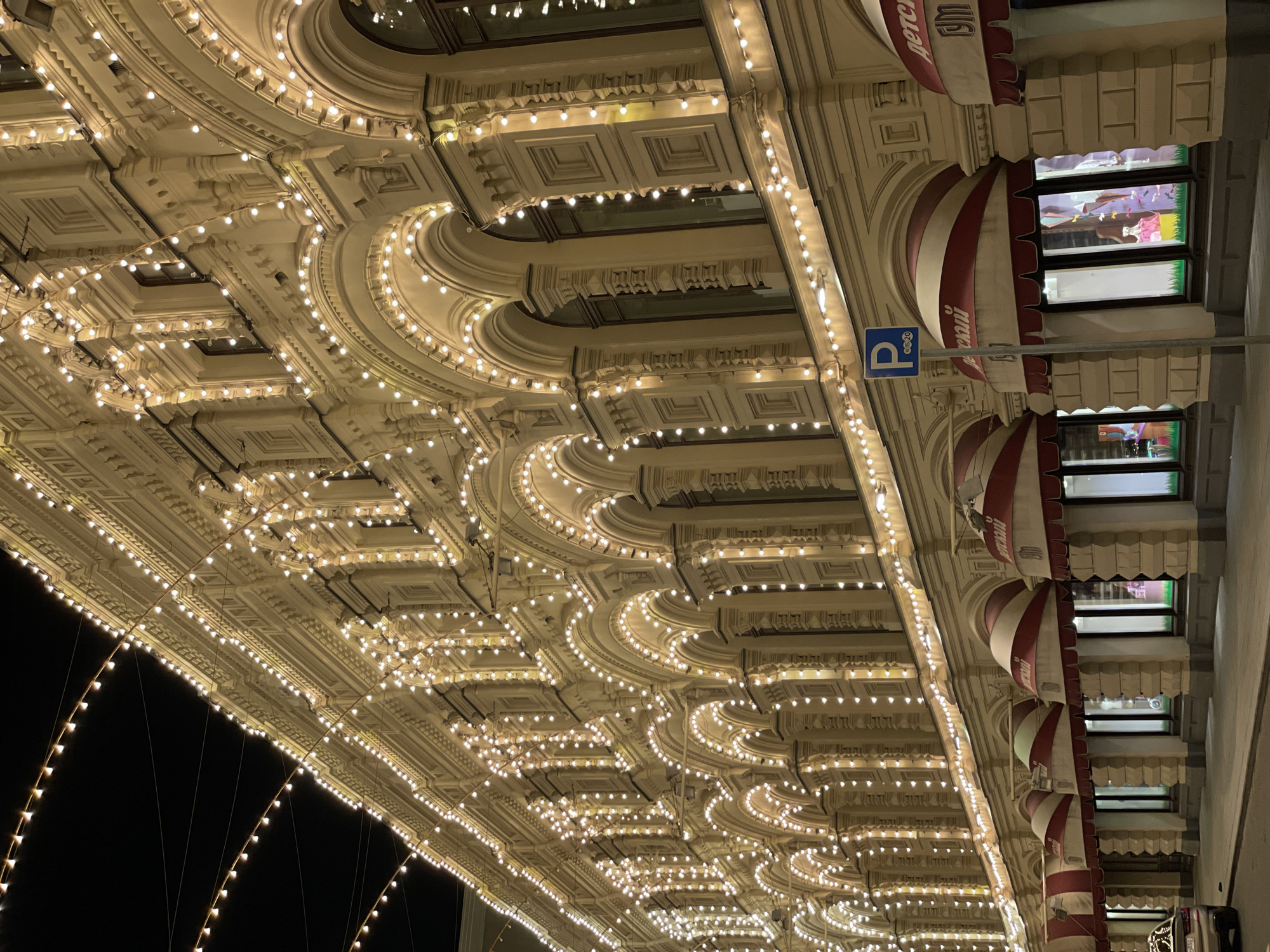 Captured from a low-angle perspective during the night, the image showcases the intricate architectural details of the GUM shopping center in Moscow, Russia. The cream-colored facade is adorned with rows of arched windows, red-and-white striped awnings, and strings of lights that highlight the building's ornamental features. The composition is oriented vertically, with the lower portion of the frame filled by the black night sky.