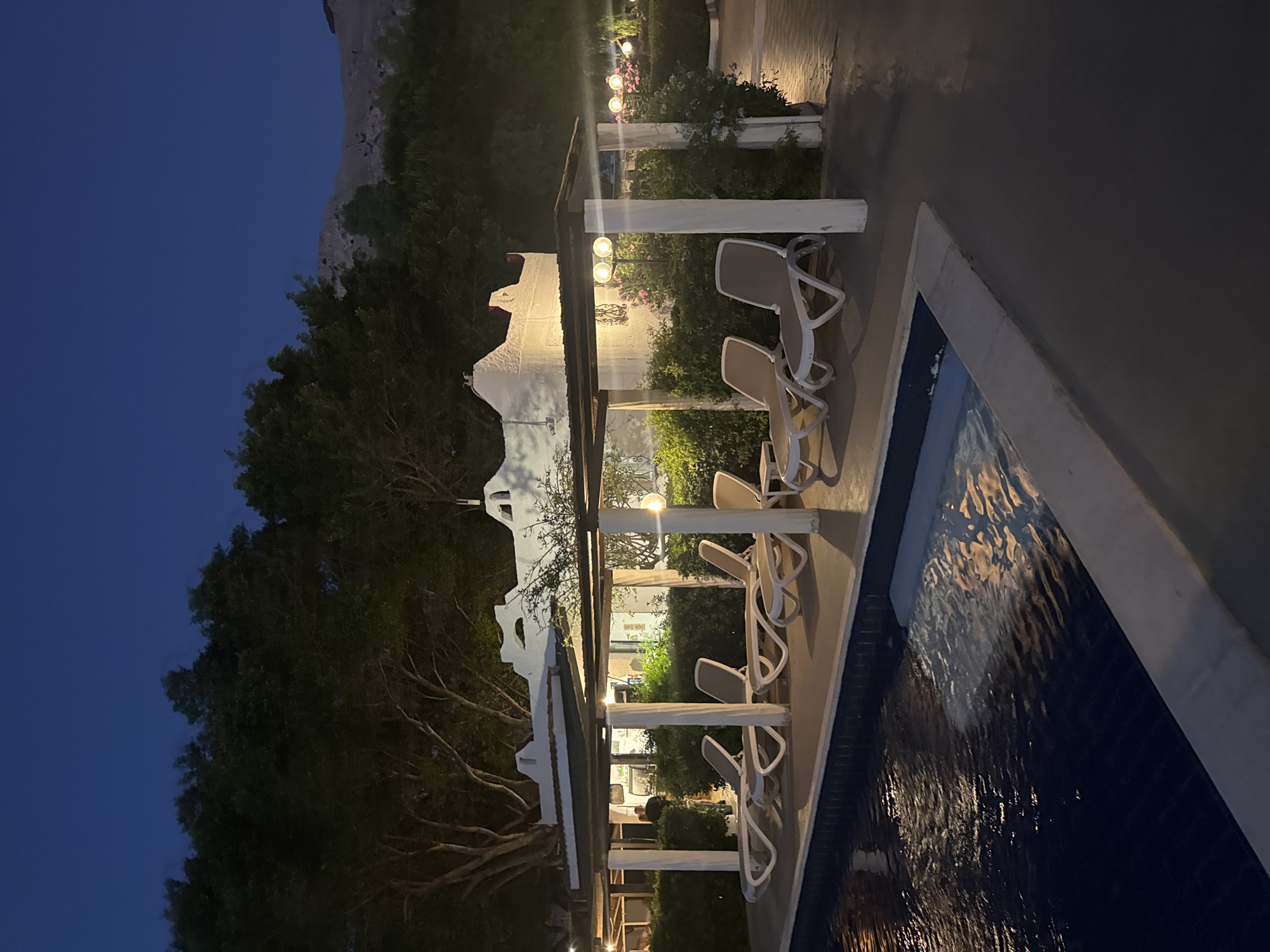 A nighttime view of a resort swimming pool area. In the foreground, a dark blue swimming pool with steps leading into it. The water reflects lights, creating a rippled pattern on the surface. Adjacent to the pool is a concrete patio with several white lounge chairs arranged under a covered walkway. The walkway has a dark roof supported by white pillars, and it extends along the side of the pool. Behind the lounge chairs, a white building with a distinctive architectural style, possibly a hotel or restaurant, is visible. It has arched openings and decorative elements. Lush green bushes and trees surround the building and the patio area. In the background, a dark, mountainous landscape is silhouetted against the twilight sky. The overall atmosphere is serene and relaxing, suggesting a vacation or resort setting in Isola delle Femmine, Italy.