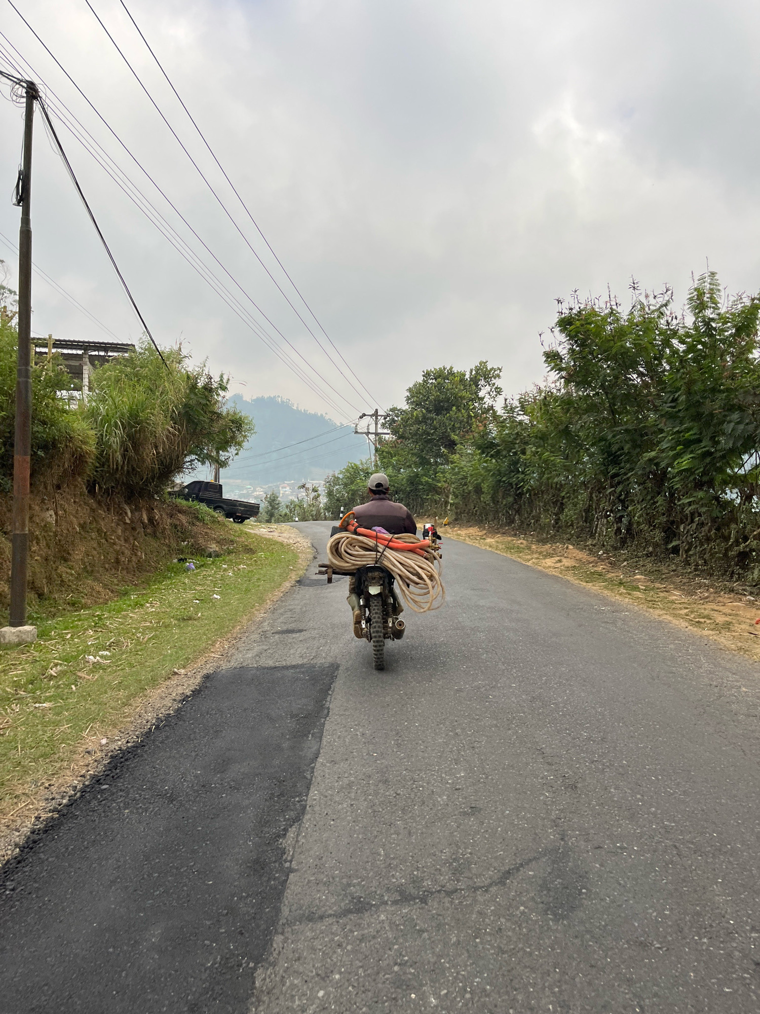 A man wearing a gray cap and a dark shirt rides a motorcycle loaded with coiled ropes and orange pipes down a paved road. The road is flanked by a grassy slope on the left and trees on the right. In the background, a town is visible in a valley with hills beyond. The sky is overcast and gray. The image appears to have been taken in Balun, Indonesia, given the context.