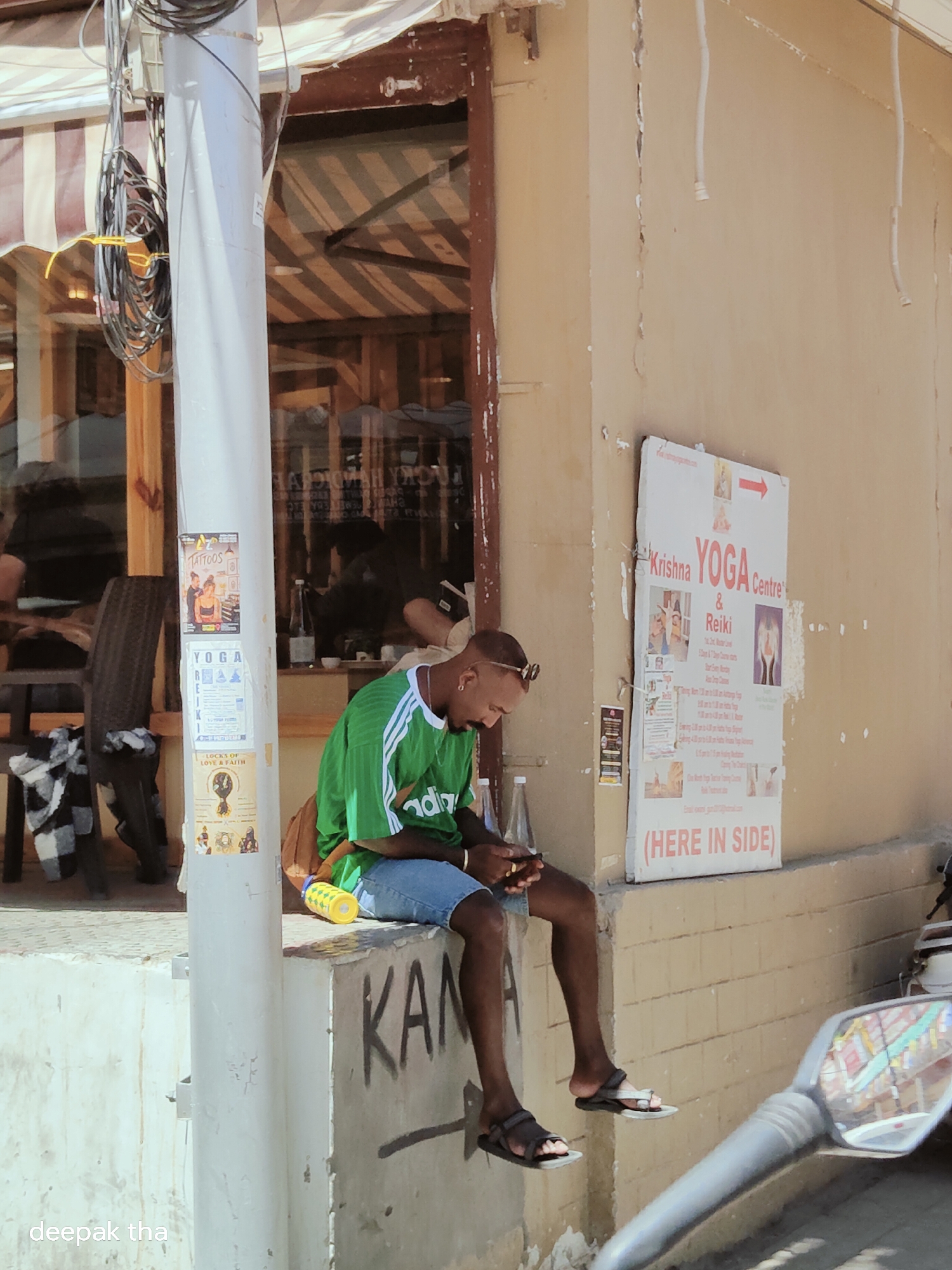 The image depicts a street scene in Leh, India, on a sunny day. A young man is seated on a concrete ledge outside a building with a sign that reads "Krishna YOGA centre & Reiki". He is wearing a green and white striped t-shirt and blue denim shorts, and he is looking down at his phone. Next to him is a brown backpack. A yellow water bottle with a green cap is also on the ledge. In the background, a window reflects the striped awning of the building. Another poster is affixed to a white pole next to the man, advertising "TATTOOS" and "YOGA". The overall impression is of a casual, outdoor urban setting.