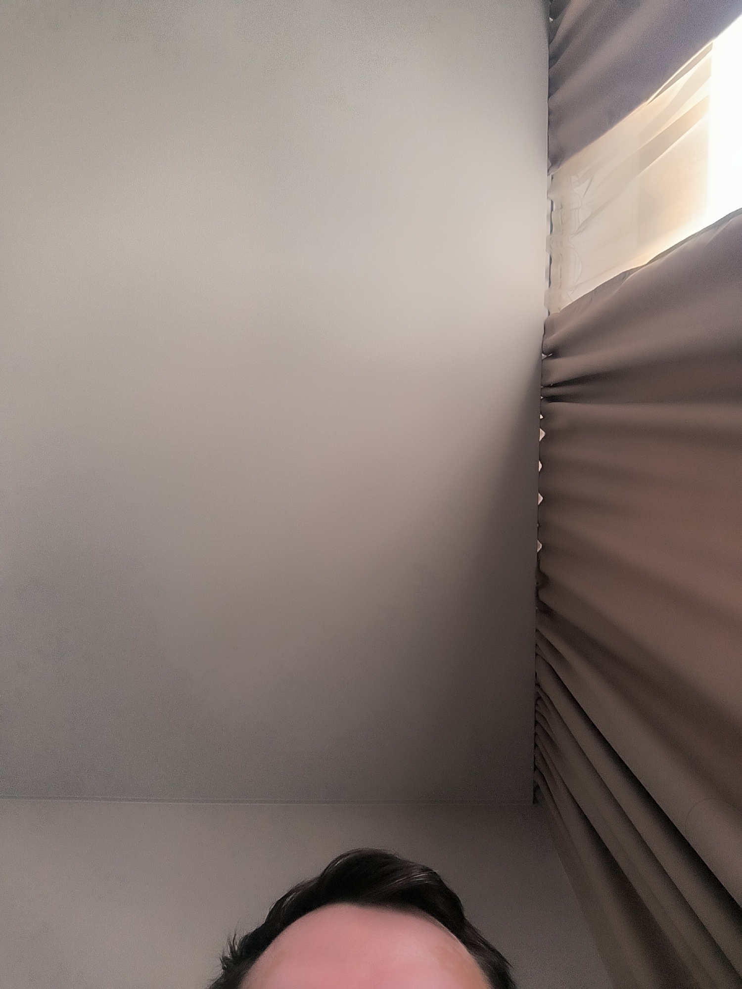 This is a close-up, indoor, eye-level shot of a person's forehead and hairline, with a plain wall and draped curtains in the background. The person's hair is dark and appears to be styled loosely. The wall is a neutral, light color and shows some subtle texture. To the right of the wall are thick, dark brown curtains, which are partially drawn to reveal a sheer, lighter curtain behind them, and a source of bright light, suggesting a window. The scene is sparsely furnished and appears to be a room within a residential or hotel setting. The location is not specified within Kovrov, Russia.