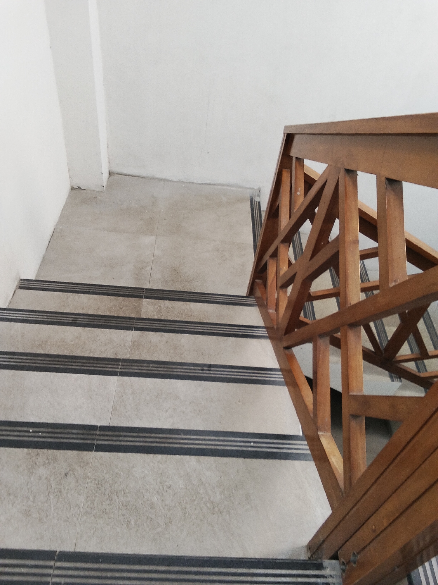 The image shows a perspective view of an interior staircase with light-colored tiled steps and dark, ribbed stair nosings. A wooden handrail with a geometric lattice design runs along the right side of the stairs. The walls and ceiling are painted white. The scene appears to be in the city of Paguyangan, Indonesia.