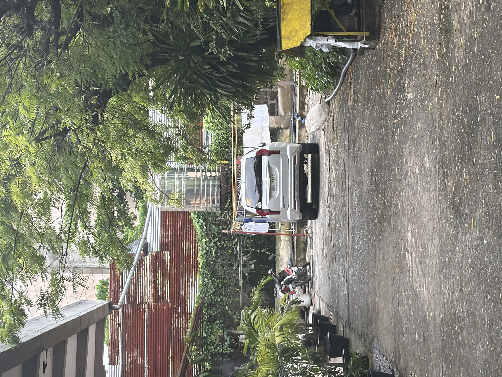 The image shows a residential scene in Davao City, Philippines. A white SUV is parked in a narrow driveway or lane, with the license plate "NHN 6760" visible. To the right of the SUV is a section of concrete pavement or a road surface. A motorcycle is parked near the SUV. A clothesline with some light-colored clothing hanging on it is also present. The surrounding area is characterized by greenery, including trees and plants. A corrugated metal fence or structure is on the left, and there are several black chairs next to some small potted plants. The overall scene is typical of a residential neighborhood in a tropical environment.