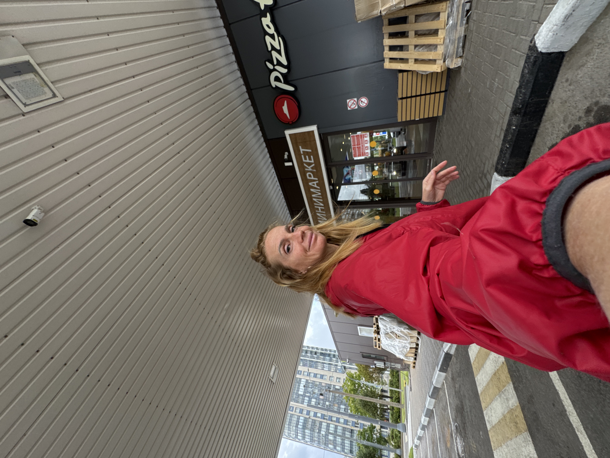 A woman wearing a bright red jacket is standing in front of a Pizza Hut and a minimarket in Saint Petersburg, Russia. She is looking towards the camera with a slight smile. The background shows a modern building, trees, and a parking lot with a pedestrian crossing. The Pizza Hut sign is clearly visible, as is the minimarket sign. There are also some wooden pallets stacked outside the minimarket. The ceiling above is made of white corrugated metal, with a security camera and a light fixture visible. The weather appears to be overcast.