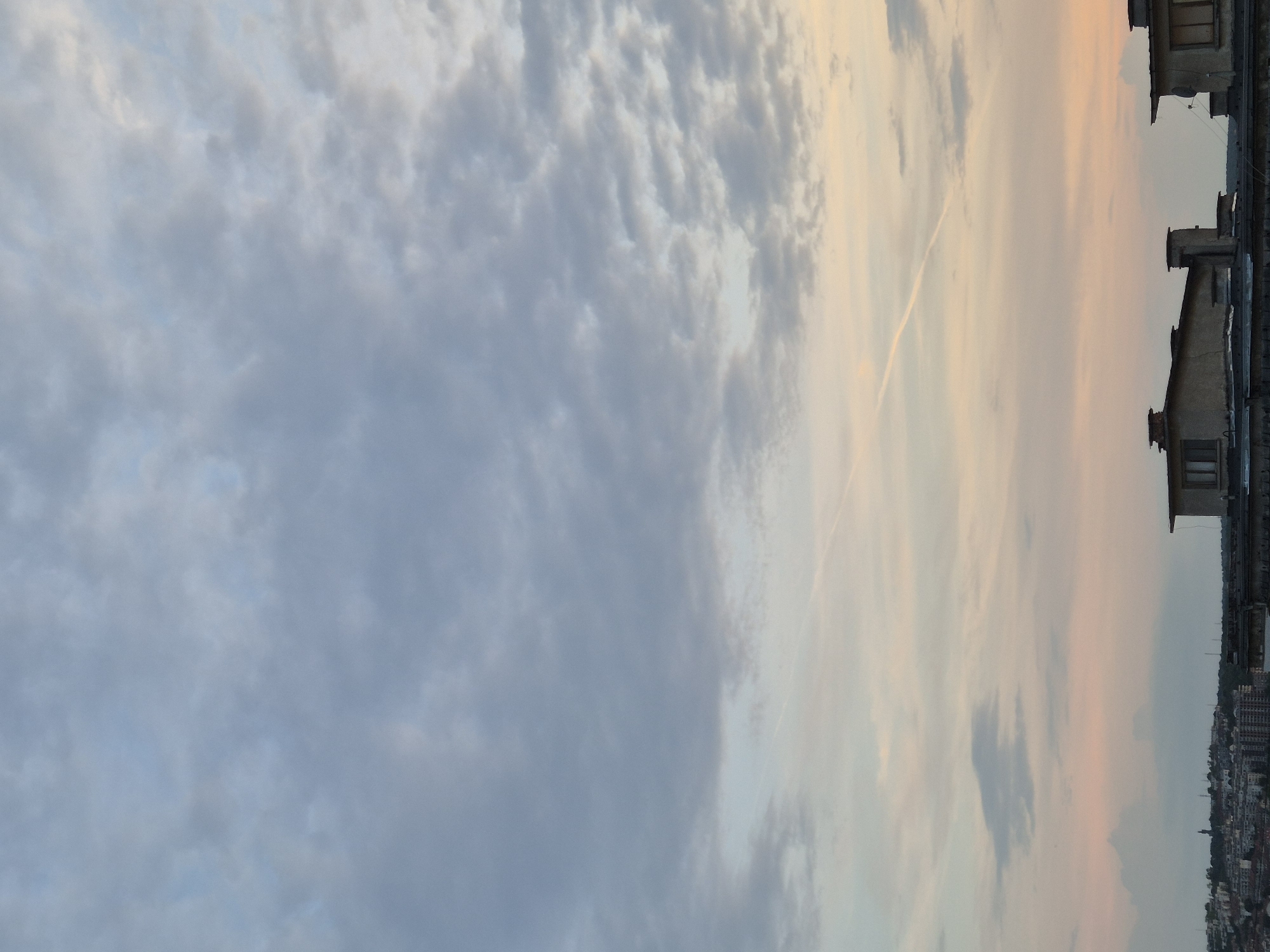 An elevated, expansive view of an urban landscape is presented at either dawn or dusk, with the specific city unknown. The upper two-thirds of the image depict a sky transitioning from an overcast, grey-blue with diffuse clouds at the top to a lighter, softer grey-pink and pale yellow near the horizon. A distinct, straight white contrail from an aircraft diagonally traverses the mid-section of the sky, extending from the upper-left towards the lower-right.
In the lower third of the image, the rooftops of several multi-story buildings dominate the immediate foreground. These structures feature flat or low-pitched roofs, multiple chimneys, and visible windows, suggesting an older architectural style, possibly concrete or stucco. A satellite dish is affixed to the rightmost foreground building. Below these prominent rooftops, the midground reveals a dense urban sprawl characterized by numerous clustered buildings of varying heights and designs, indicative of a city. On the far left, a distant hill or elevated area is visible, topped by a tall, slender structure, potentially a monument, statue, or communication tower, silhouetted against the lighter sky. No individuals are visible within the frame, and no specific actions are discernible. The overall lighting is soft and diffused, characteristic of twilight conditions.