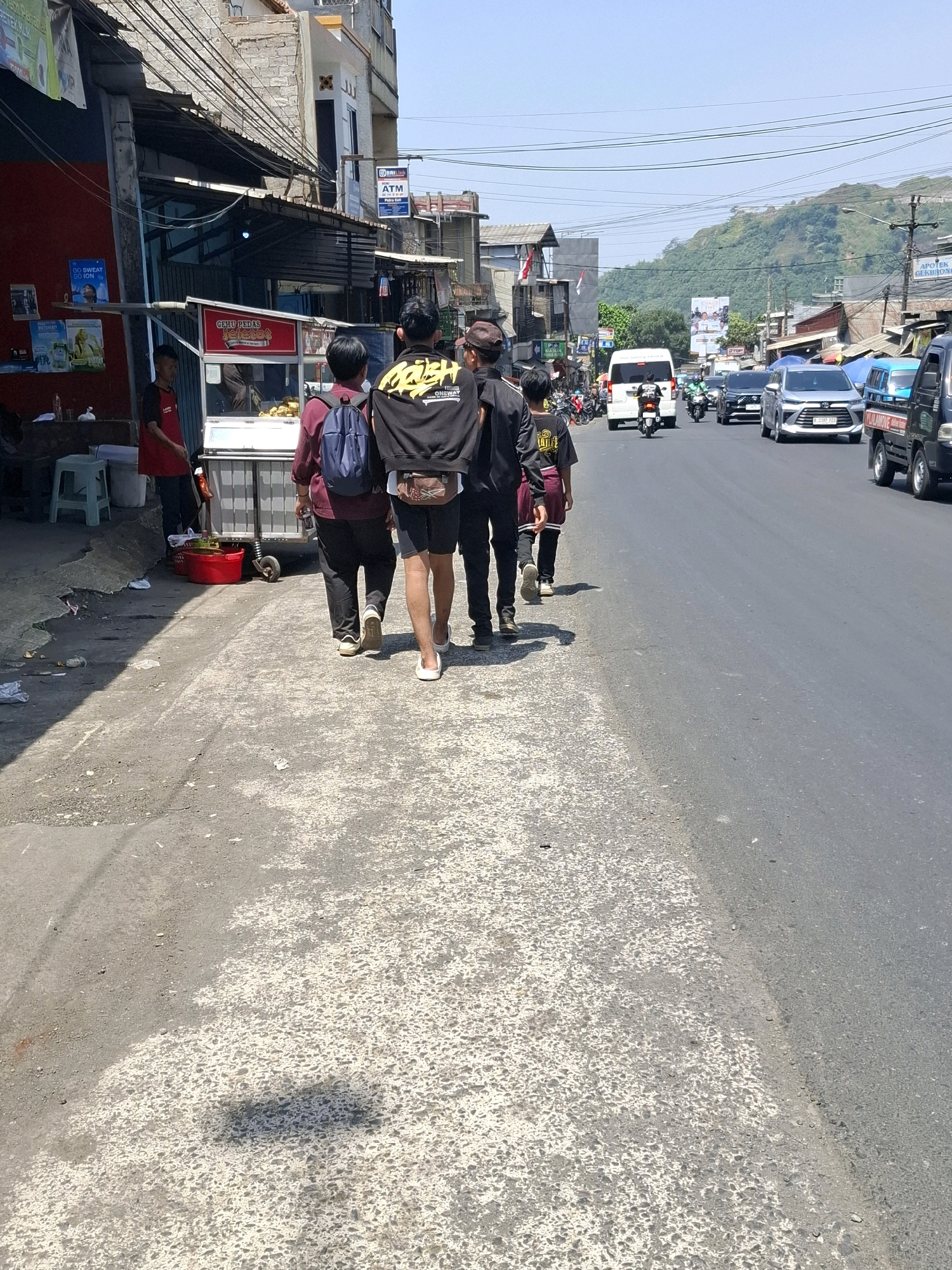 In Cikahuripan, Indonesia, on a sunny day, four young individuals walk away from the camera on a cracked asphalt sidewalk bordering a busy road. The group consists of three males wearing black shirts and one male in a maroon shirt with a blue backpack. The male in the center of the group wears a black hoodie with yellow lettering. They are walking towards a street with several vehicles, including cars, motorcycles, and a white van. On the left side of the sidewalk, there is a food stall with a red cart and a person in a red shirt standing behind it. Buildings line the street on the left, and a green, tree-covered hill is visible in the background. Power lines crisscross above the scene.