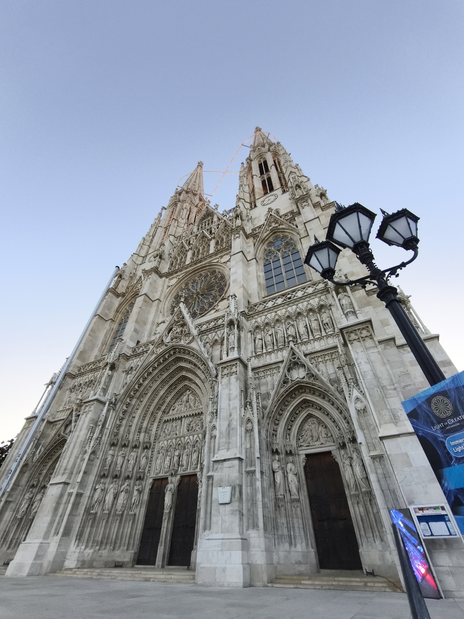 This is a low-angle, wide-shot photograph of the Votivkirche in Vienna, Austria, under a clear, pale blue sky. The Gothic Revival church, constructed of light-colored stone, dominates the frame, with its intricate facade, spires, and detailed sculptures visible. The perspective emphasizes the grandeur and height of the building. In the foreground, to the right, a black, ornate, three-headed lamppost stands. Adjacent to the lamppost, a tall, angled blue sign with white text is partially visible, and below it, a smaller display board shows colorful light projections. The ground in front of the church appears to be paved. The image captures the exterior of a significant architectural landmark in Vienna.