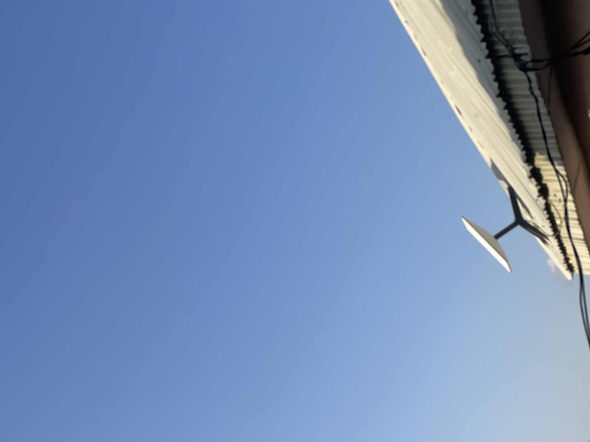 A satellite dish, likely a Starlink dish, is mounted on a corrugated metal roof. The dish is white and has a rectangular shape with rounded corners, angled towards the sky. The roof appears weathered, with some signs of rust and discoloration. Wires are visible running along the edge of the roof, connected to some unseen infrastructure. The scene is set against a clear blue sky, indicating daytime. The image is taken from a low angle, looking up towards the roof and the sky. The location is Unknown, Nigeria.
