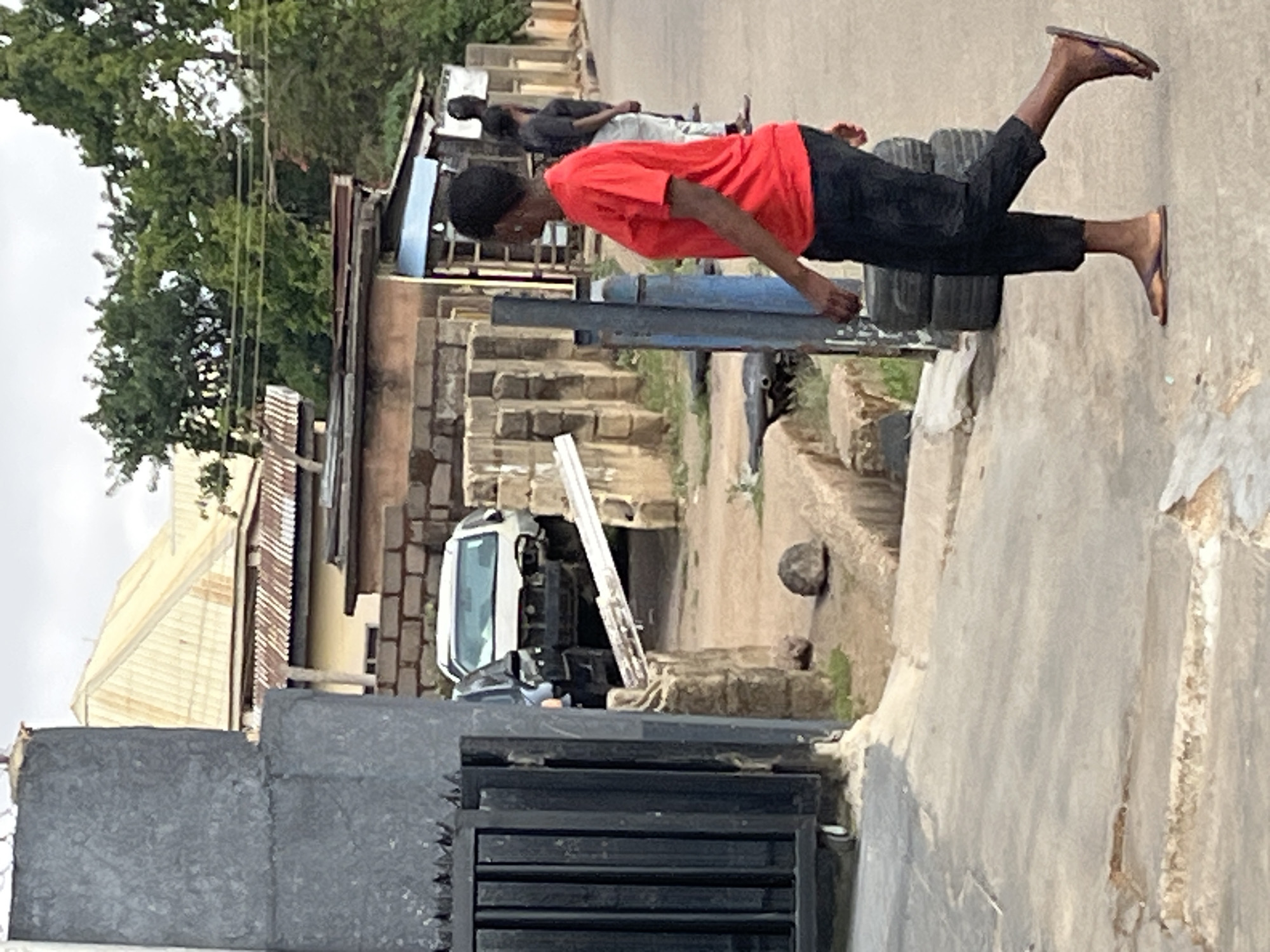 The image depicts a young person in a red shirt and dark pants doing push-ups on a stack of tires on a street in Jos, Nigeria. They are wearing flip-flops. Another person is lying on a nearby stack of tires. The street scene includes buildings, trees, a white vehicle, and a partly-constructed cement structure.