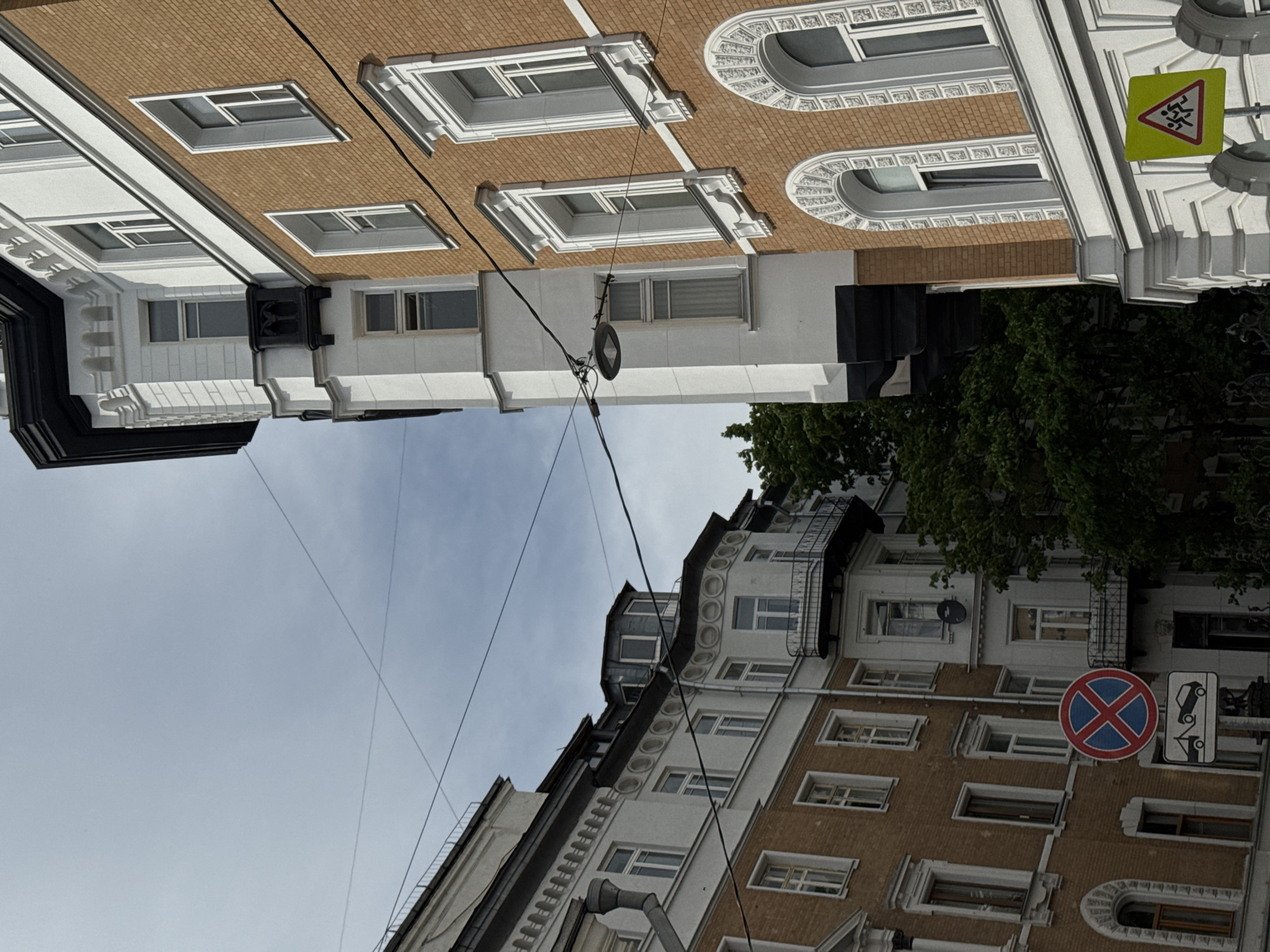 The image depicts an urban scene in Moscow, Russia, featuring buildings with architectural details like decorative windows and cornices. Street signs, including a "no parking" sign and a pedestrian crossing sign, are visible, indicating a city street. Overhead, power lines crisscross against the sky. The architectural style and street signs suggest a European urban environment. The sky is cloudy, contributing to the overall daylight setting.