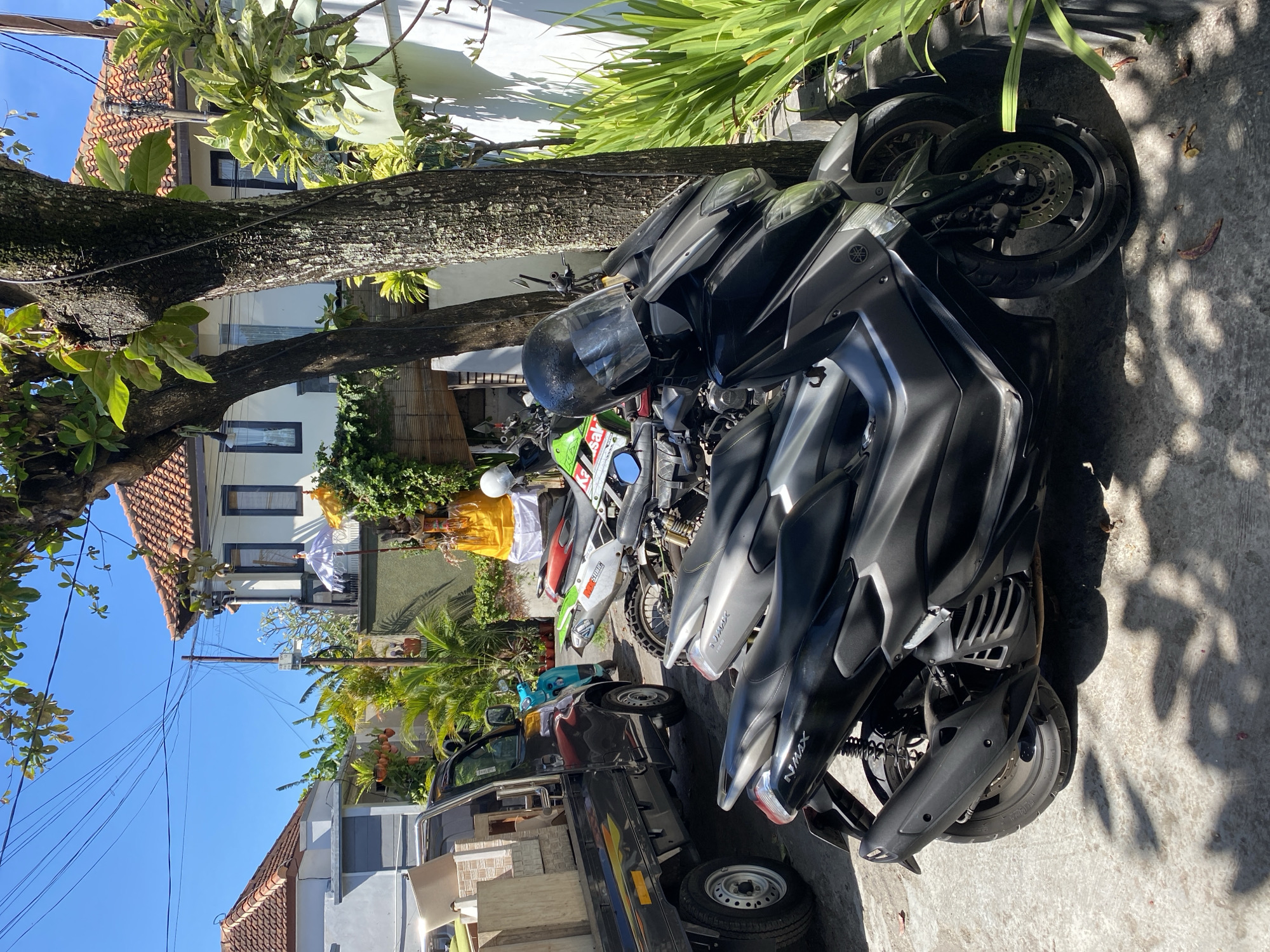 In Denpasar Barat, Indonesia, a sunny day is depicted with a street scene featuring multiple motorcycles parked under trees. In the foreground, two dark-colored scooters are parked side-by-side, with the closest one being a Yamaha NMAX. Behind them, a green and white dirt bike is visible, followed by another motorcycle. A small black utility truck is parked to the left, loaded with light-colored boxes. In the background, traditional Balinese architecture with red-tiled roofs and white walls is visible, along with lush green vegetation and palm trees. A religious offering, possibly a Balinese ceremony, is set up with a yellow cloth and a white umbrella. The environment appears to be residential or commercial, with power lines visible overhead. The overall impression is of a typical day in a tropical urban environment.