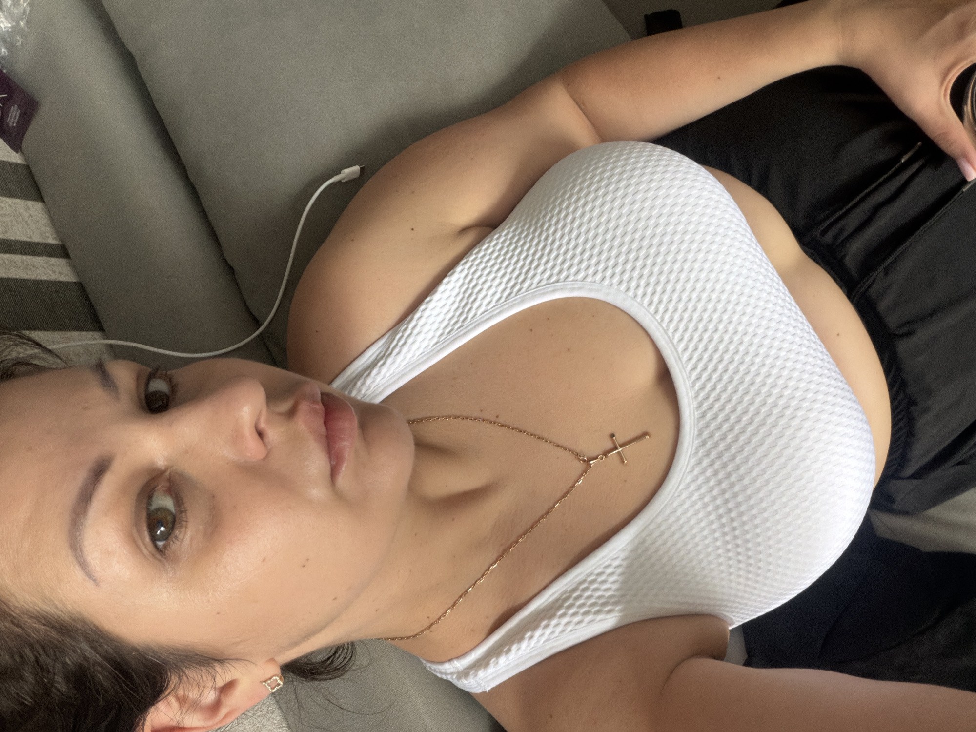 A woman is lying on a grey couch, looking up at the camera. She is wearing a white sports bra and black athletic shorts. She has dark hair pulled back and is wearing a gold necklace with a cross pendant. A white cable is visible behind her on the couch. The image does not provide any information about the location in the city of Sochi, Russia.