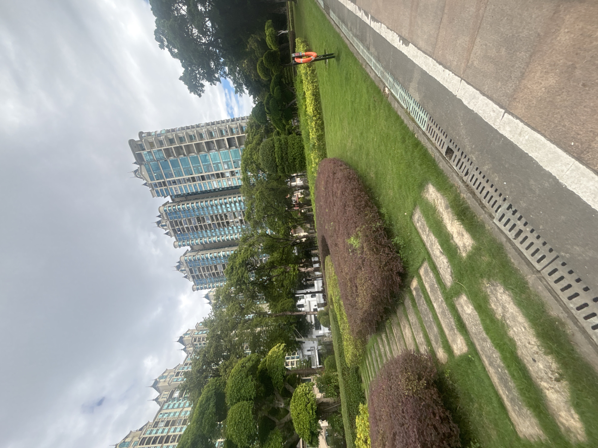 The image depicts a park area in the city of Nanhai District, China, featuring a meticulously landscaped garden with a variety of flora. Tall residential buildings with distinctive architectural styles, characterized by multiple stories, balconies, and ornate rooftops, dominate the background. The foreground showcases lush green grass interspersed with stone pathways and manicured bushes in hues of green and deep red. A drainage grate runs along the edge of an asphalt pathway, marked by a white line, separating the garden from the path. A red lifebuoy is mounted on a pole within the park. The sky above is overcast with scattered clouds, suggesting a temperate climate. There are no people visible in the frame.
