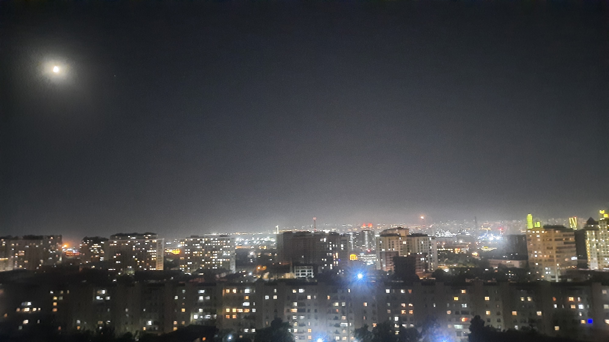 The image is a nighttime panoramic view of a city skyline under a dark, hazy sky. The moon is visible in the upper left quadrant of the image, casting a soft glow. The city is illuminated by numerous lights from buildings and streetlights, creating a sprawling urban landscape. The buildings appear to be a mix of residential and commercial structures, with varying heights and architectural styles. The overall impression is one of a busy and active city at night. The specific location is unknown.