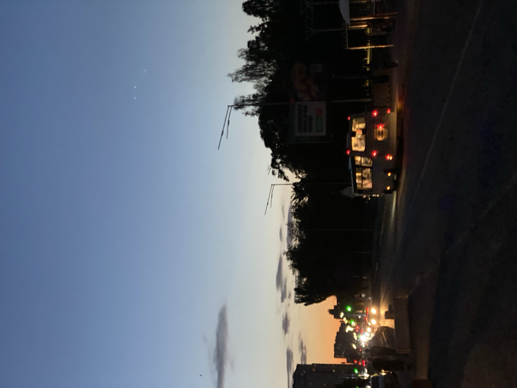 An outdoor, ground-level, eye-level shot captures a street scene at dusk in an unspecified urban location. The sky transitions from a pale orange on the horizon to a deep blue overhead, with faint clouds visible. Three distant lights, possibly stars or celestial objects, are visible in the upper right portion of the sky. Silhouetted trees and buildings form a dark backdrop along the right and left sides of the image, respectively.
A street with a traffic lane runs through the center of the frame, dimly lit by the headlights of approaching vehicles and streetlights. On the left side of the road, a line of cars with illuminated headlights and taillights are seen. Two figures, partially obscured by the low light, are walking on the left sidewalk. A bus, with its rear red taillights illuminated, is traveling down the street towards the right. There are streetlights and signs, including a billboard, along the right side of the road. The overall atmosphere is that of a city street at the end of the day, with artificial lights beginning to dominate the fading natural light.