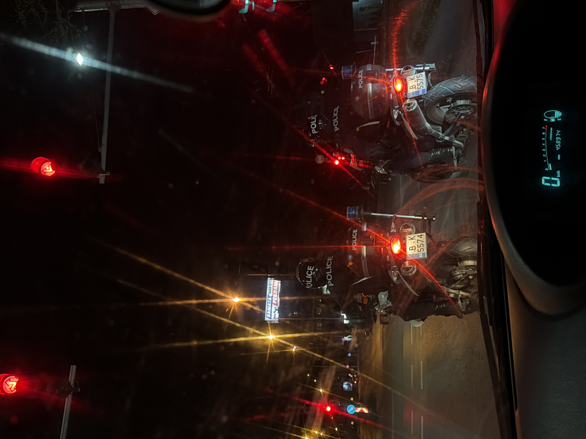 The image, taken from inside a vehicle in Varna, Bulgaria, depicts a line of stopped police motorcycles at night. The motorcycles have "POLICE" printed on the back and bear license plates with "B.K" and numbers "5574" and "5575". The car's dashboard displays "0 km/h" and an odometer reading of "458974". Traffic lights are visible, with the color red. A sign with the Cyrillic words "ВАШАТА РЕКЛАМА" ("YOUR ADVERTISEMENT") and a phone number starting with "0884" is illuminated nearby.