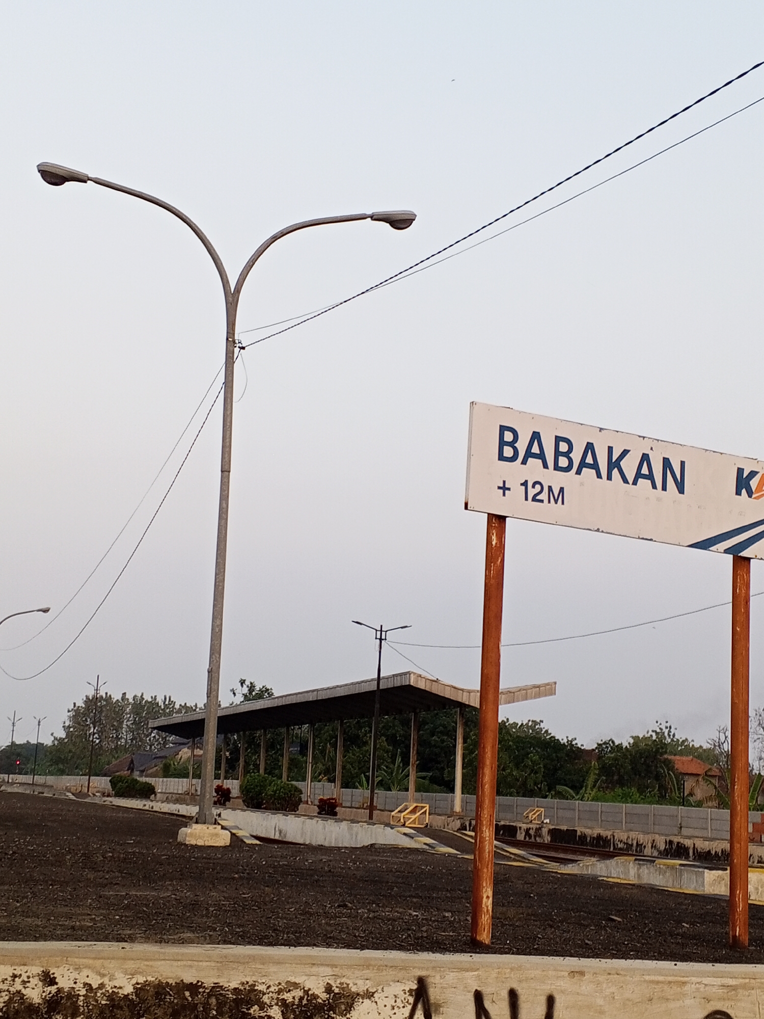 The image shows a sign that reads "BABAKAN +12M K" in front of what appears to be a train station in Pakusamben, Indonesia. The sign is white with blue lettering and is supported by two rusty orange poles. In the background, there is a covered waiting area with a dark roof, and train tracks are visible. The ground in front of the station is covered in dark, loose material, possibly coal or dirt. There are several streetlights and power lines visible in the sky, which is overcast. Trees and some buildings are visible in the distance. The overall scene suggests a functional but perhaps not highly trafficked train station, typical of a smaller town or rural area.